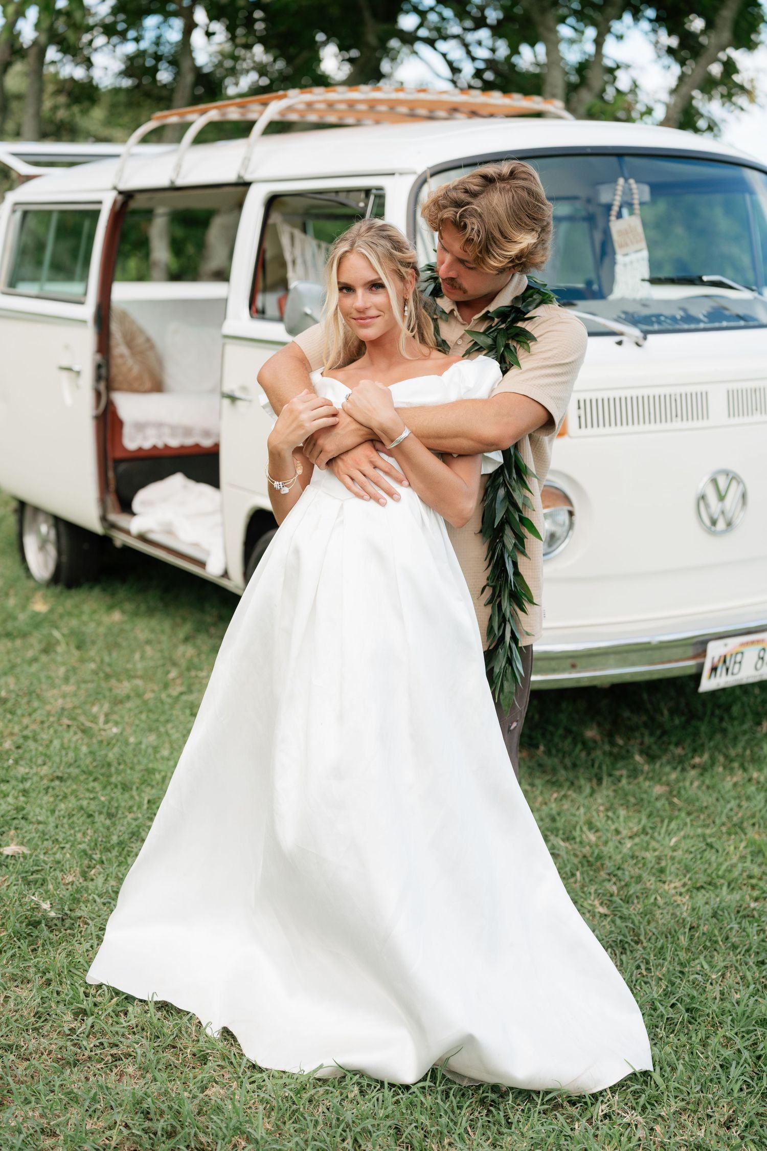 Bride and groom embrace in front of a vintage VW bus on green grass, groom wearing a tropical lei.