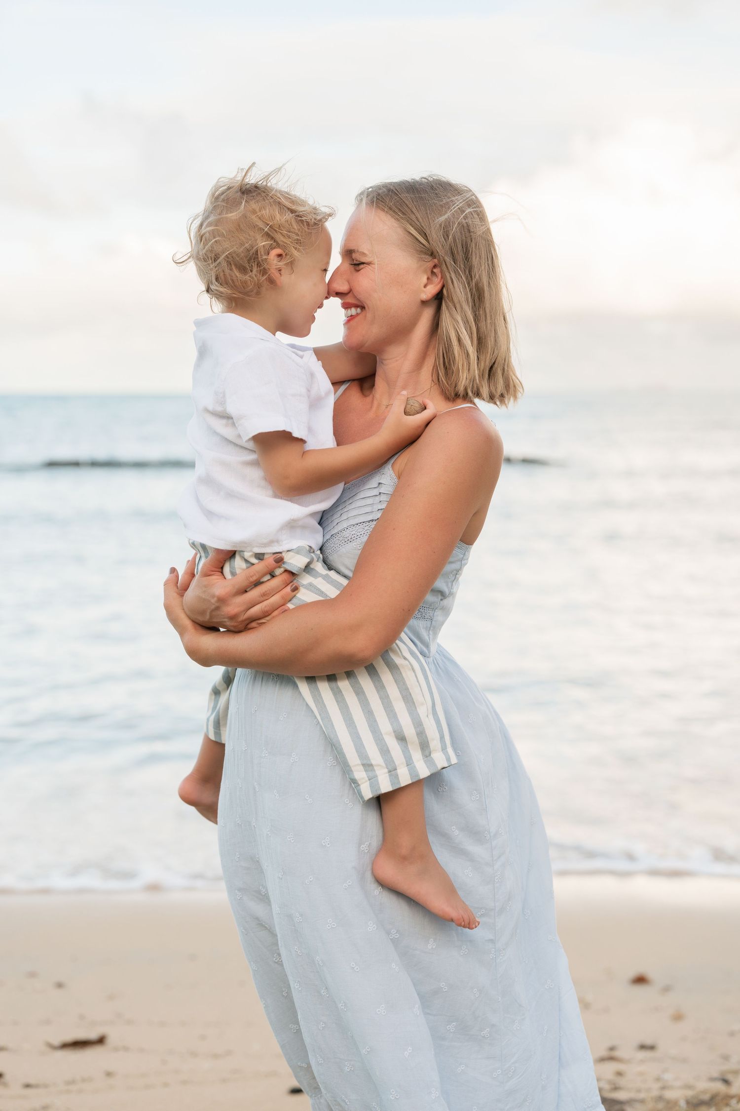 Smiling mother holds toddler nose-to-nose on a sandy beach at sunset.