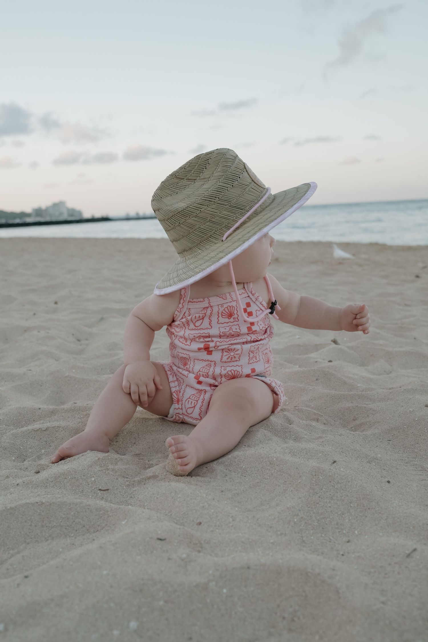 Baby in pink swimsuit and straw hat sitting on sandy beach at sunset.