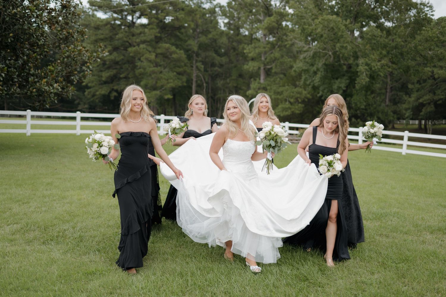 A wedding party poses in a grassy field with white fences, wearing black bridesmaid dresses and a white bridal gown.