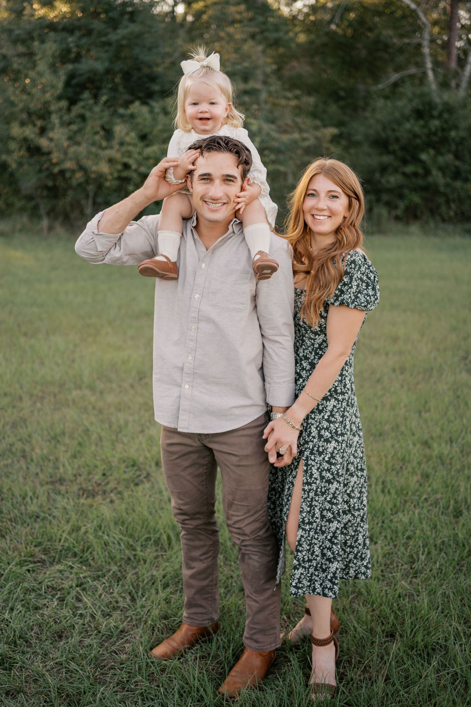 A family stands together in a grassy field at golden hour, wearing casual clothing and sharing a tender moment outdoors.