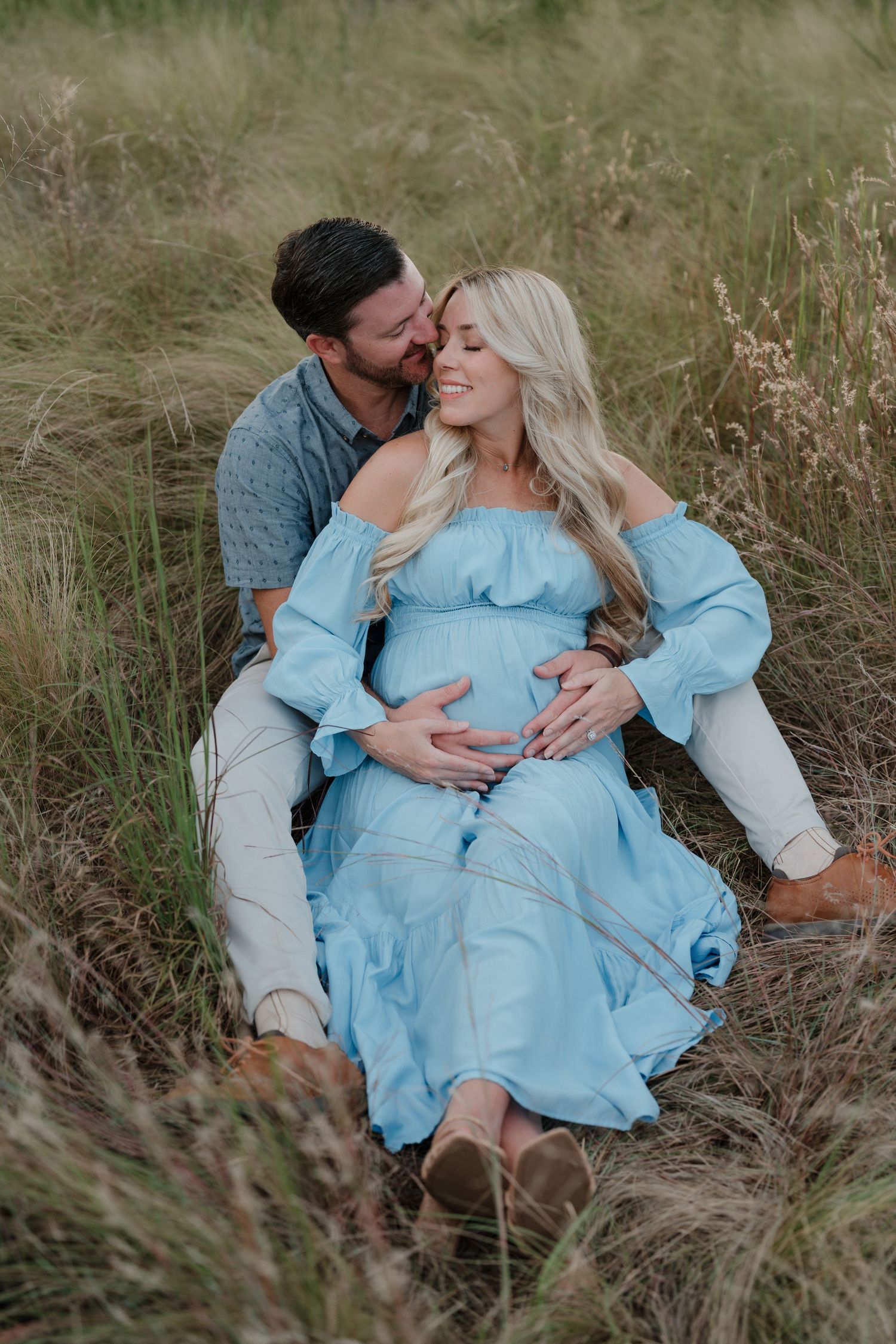 A couple shares an intimate moment in a field of tall grass as they celebrate their pregnancy in a light blue dress.