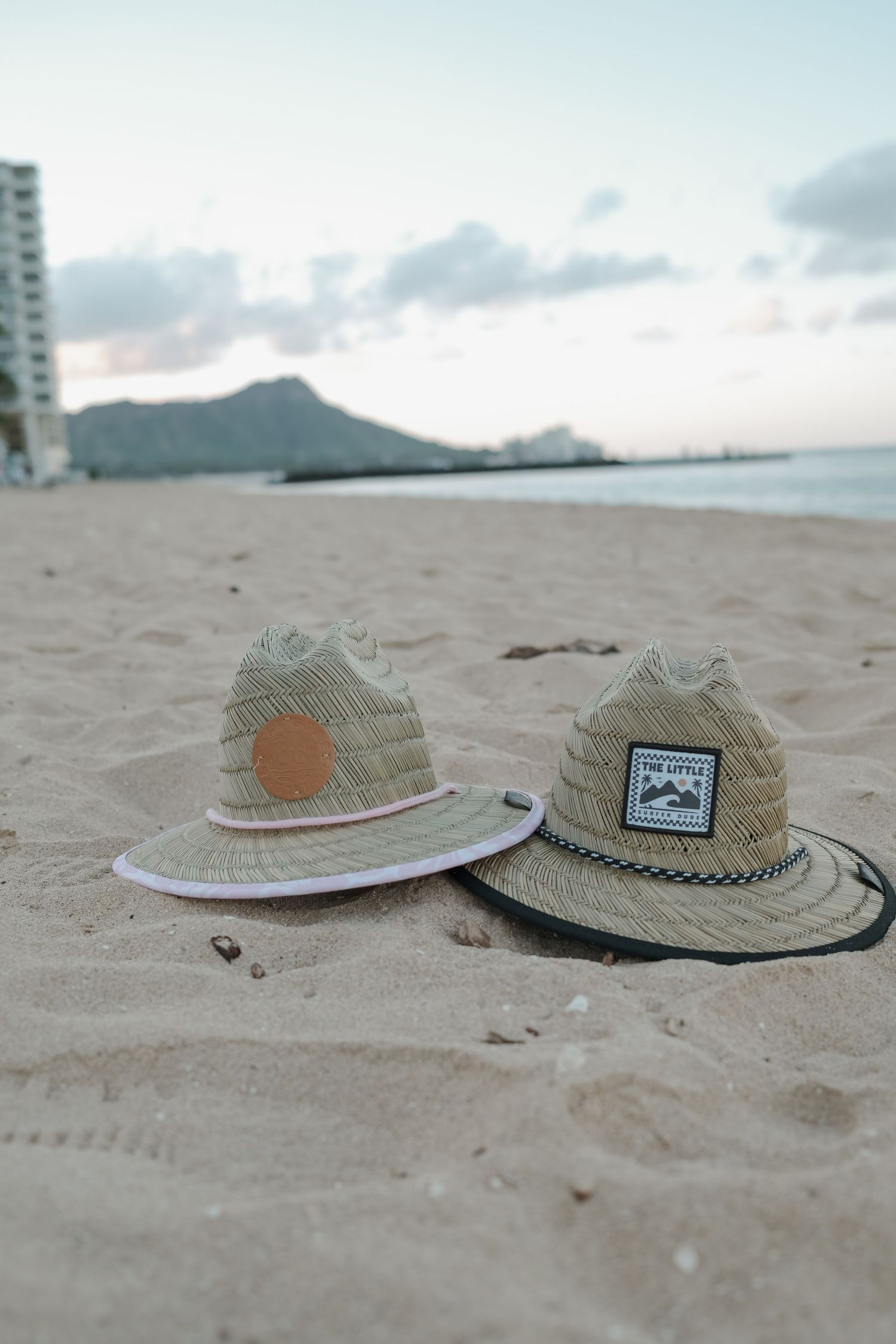 Two straw lifeguard hats resting on sandy Waikiki Beach with Diamond Head crater in the background at sunset.