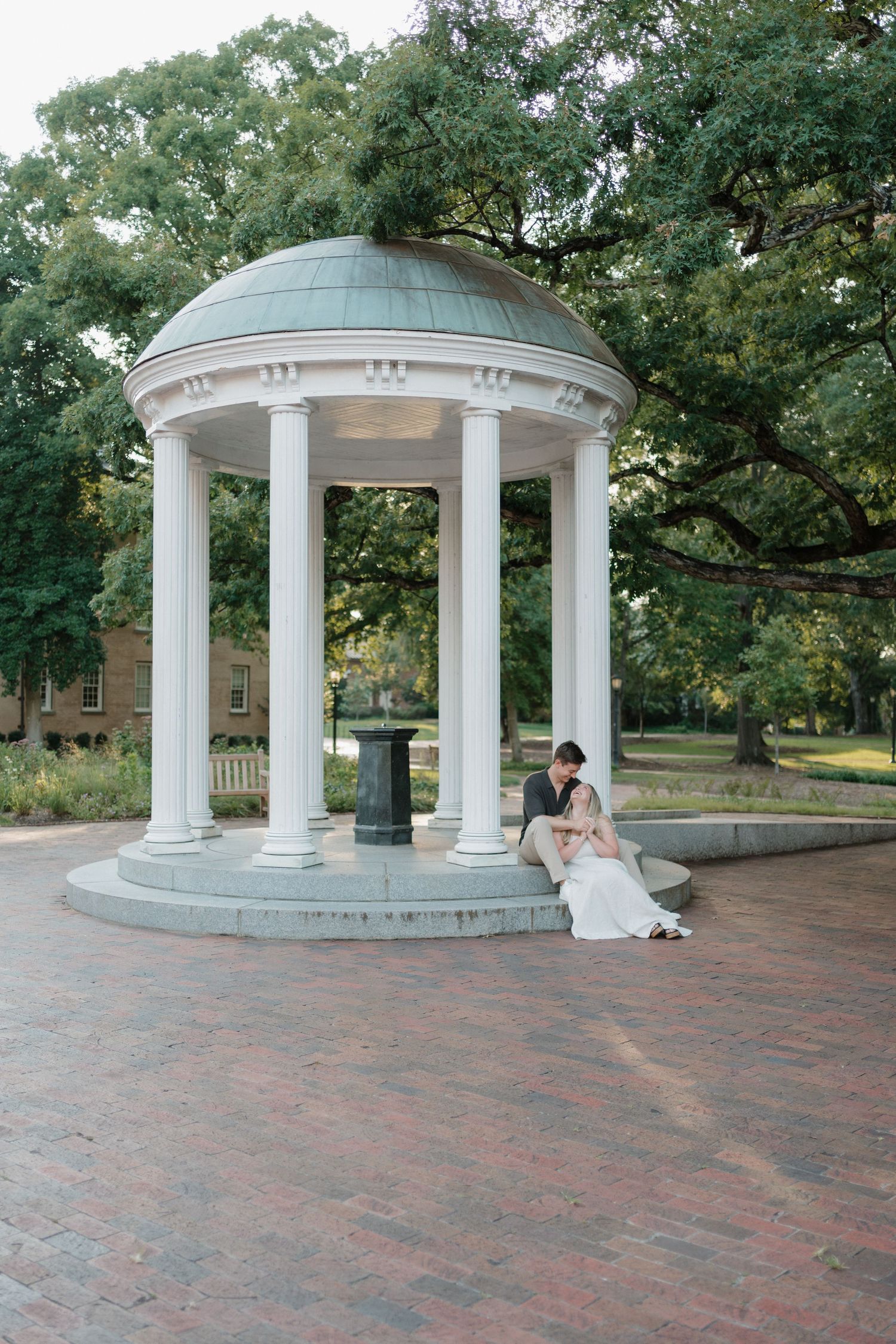 White columned gazebo with a domed roof sits on a brick patio surrounded by green trees and a historic building.