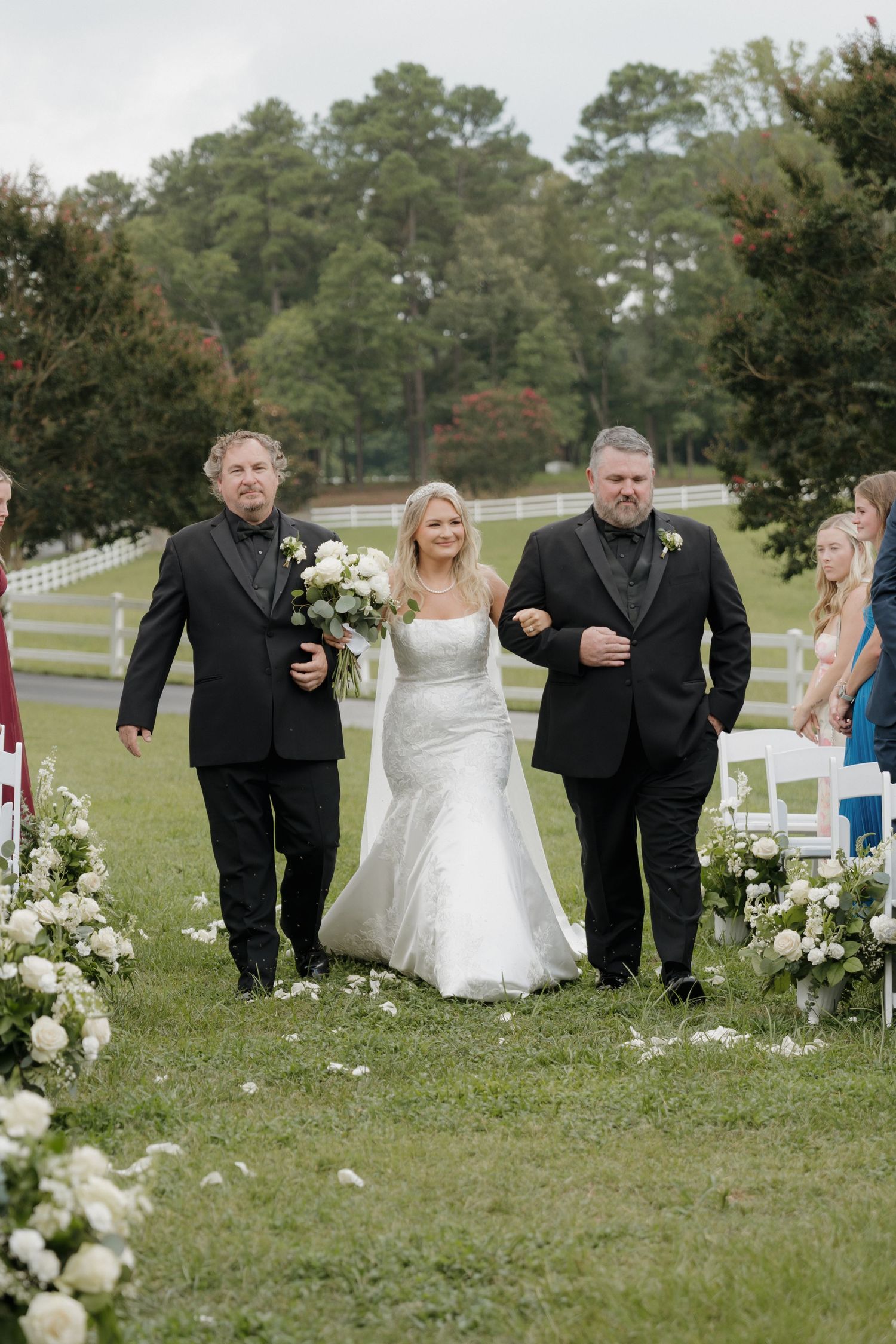 Wedding party members in black suits and white dress playfully walking through scattered flower petals on grassy lawn.