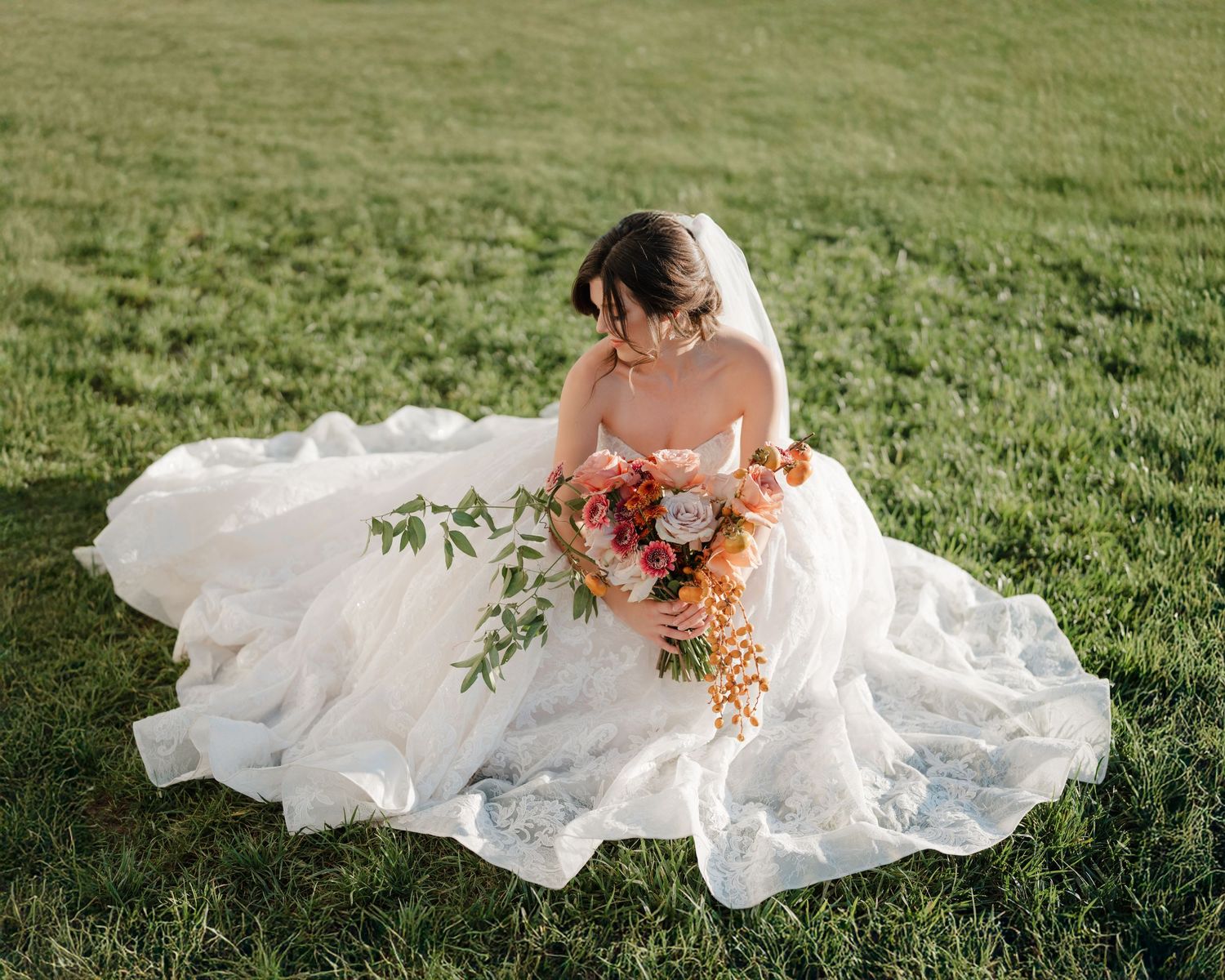 Flowing white wedding dress billows in the breeze across lush green grass as bride holds vibrant coral bouquet.