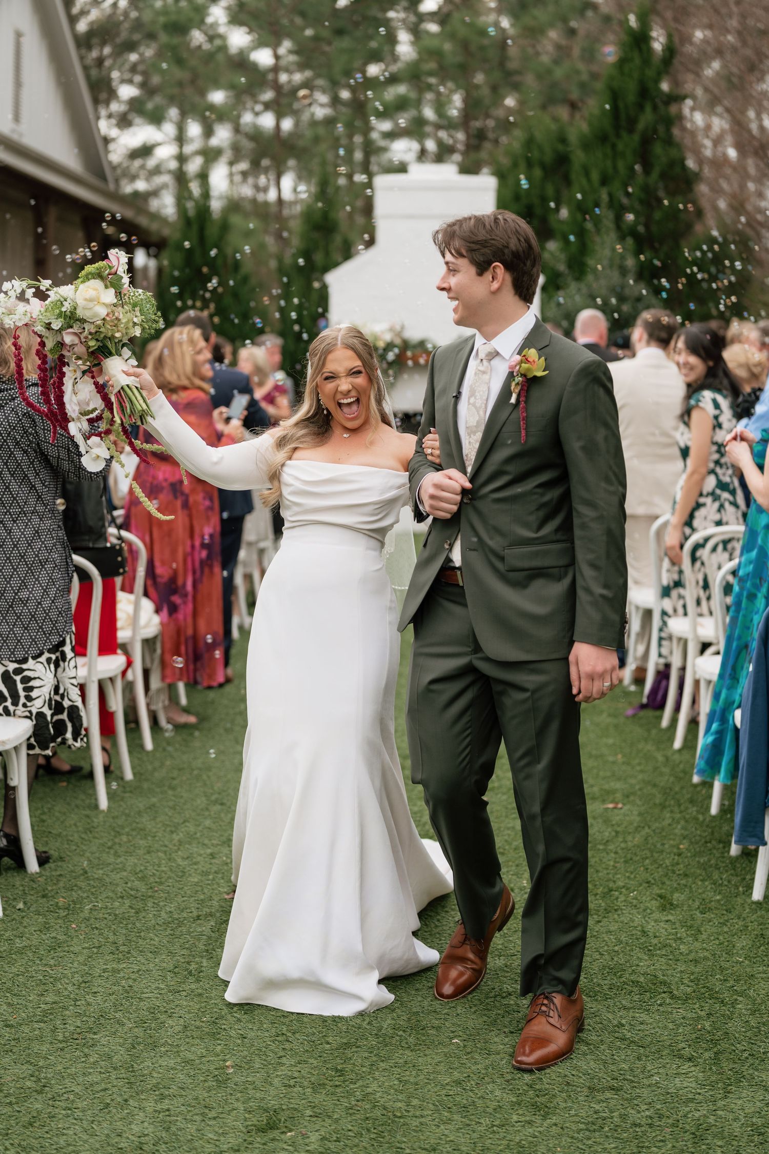 Joyful bride in white gown and groom in green suit walk down outdoor wedding aisle as guests celebrate.