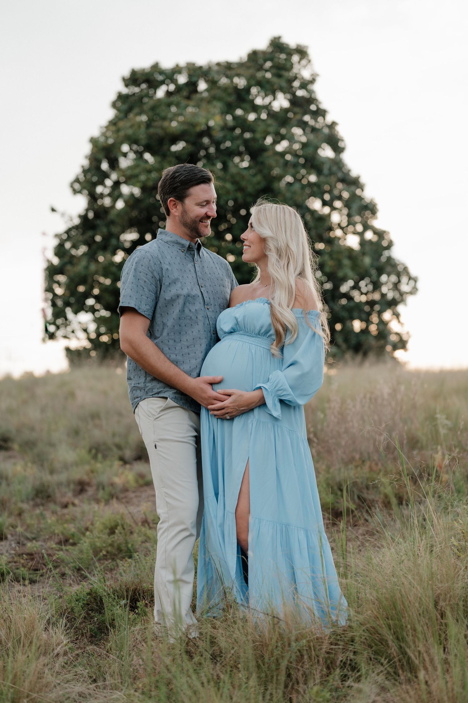 An expecting couple poses together in a field at sunset, with her wearing a flowing light blue maternity dress.