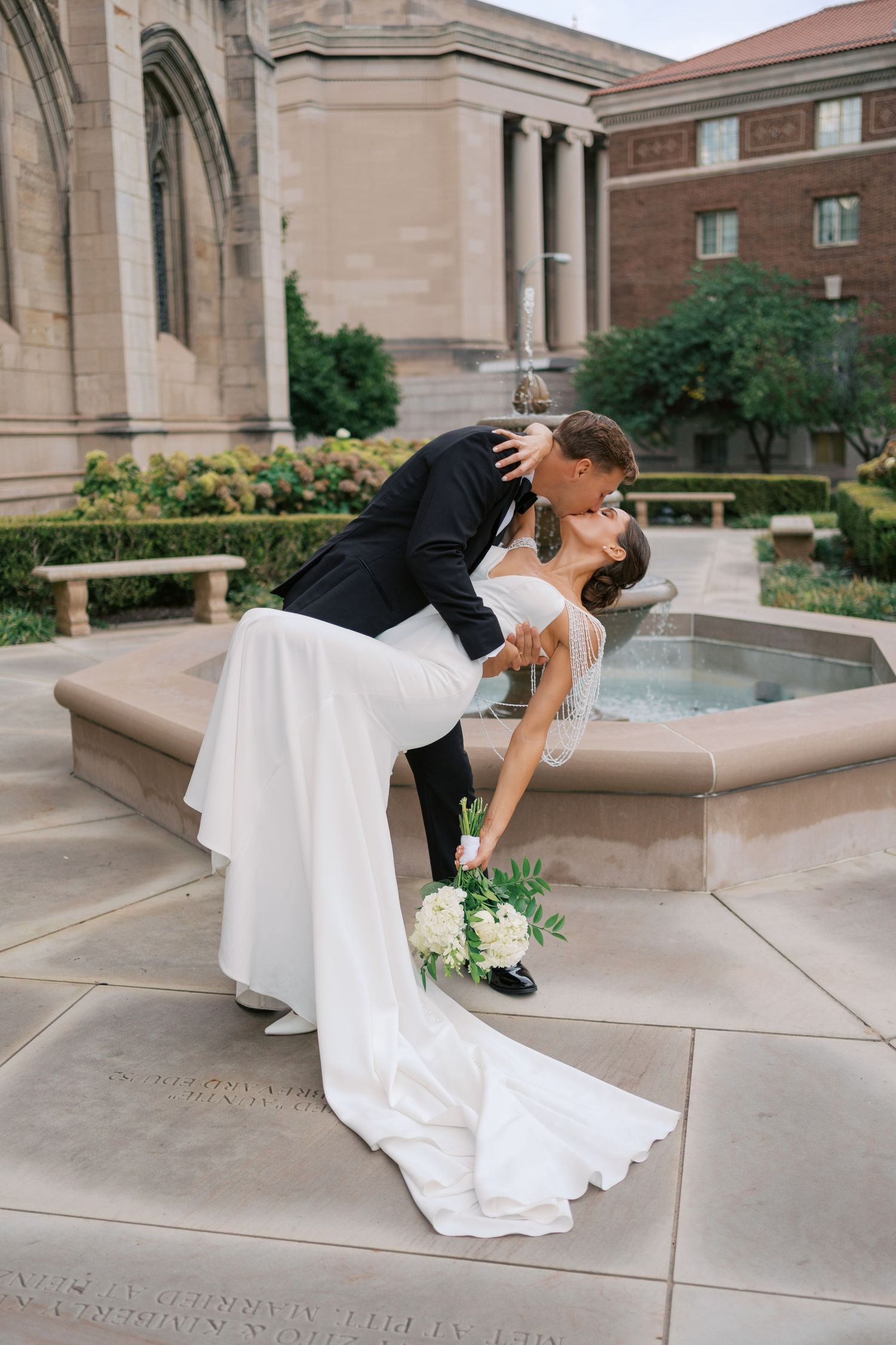 Romantic wedding couple shares intimate dips and kisses in front of an elegant stone building with fountain.