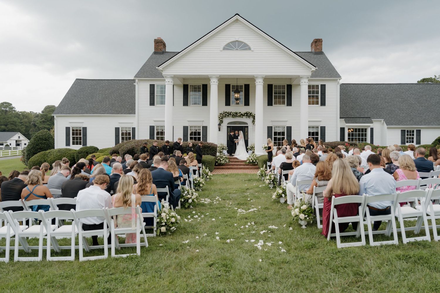 Elegant outdoor wedding ceremony setup on a lawn in front of a grand white colonial style mansion with black shutters.