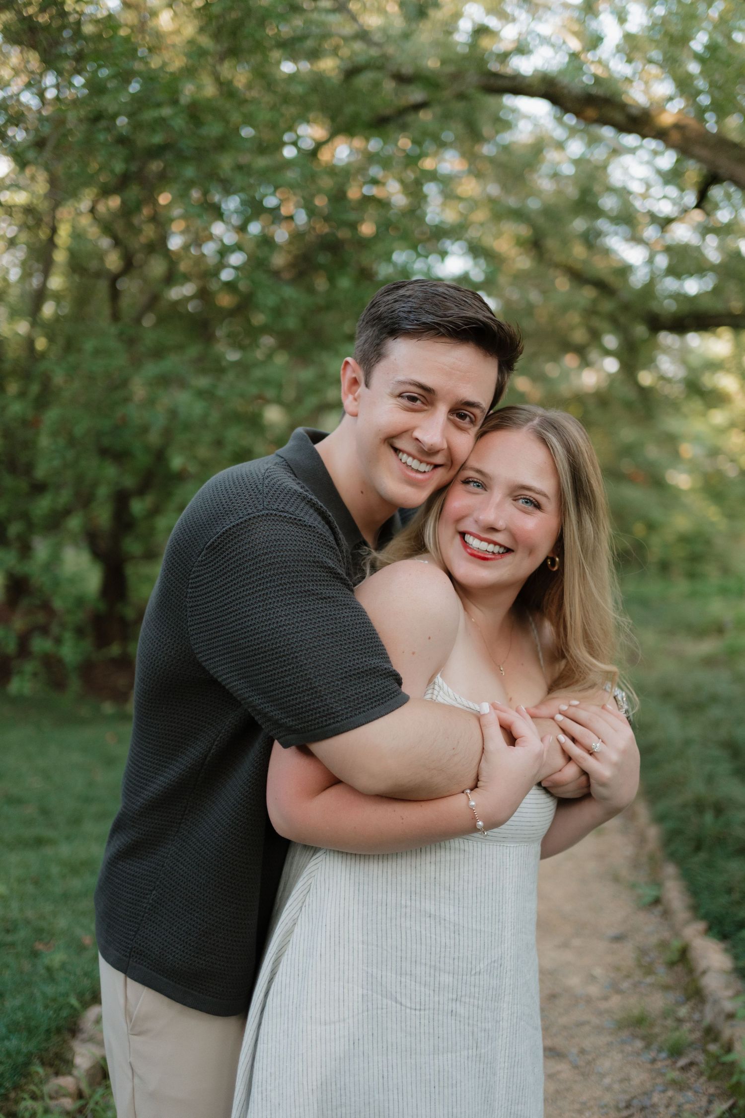 A couple shares joyful embraces and laughter while posing together on a nature path surrounded by green trees.