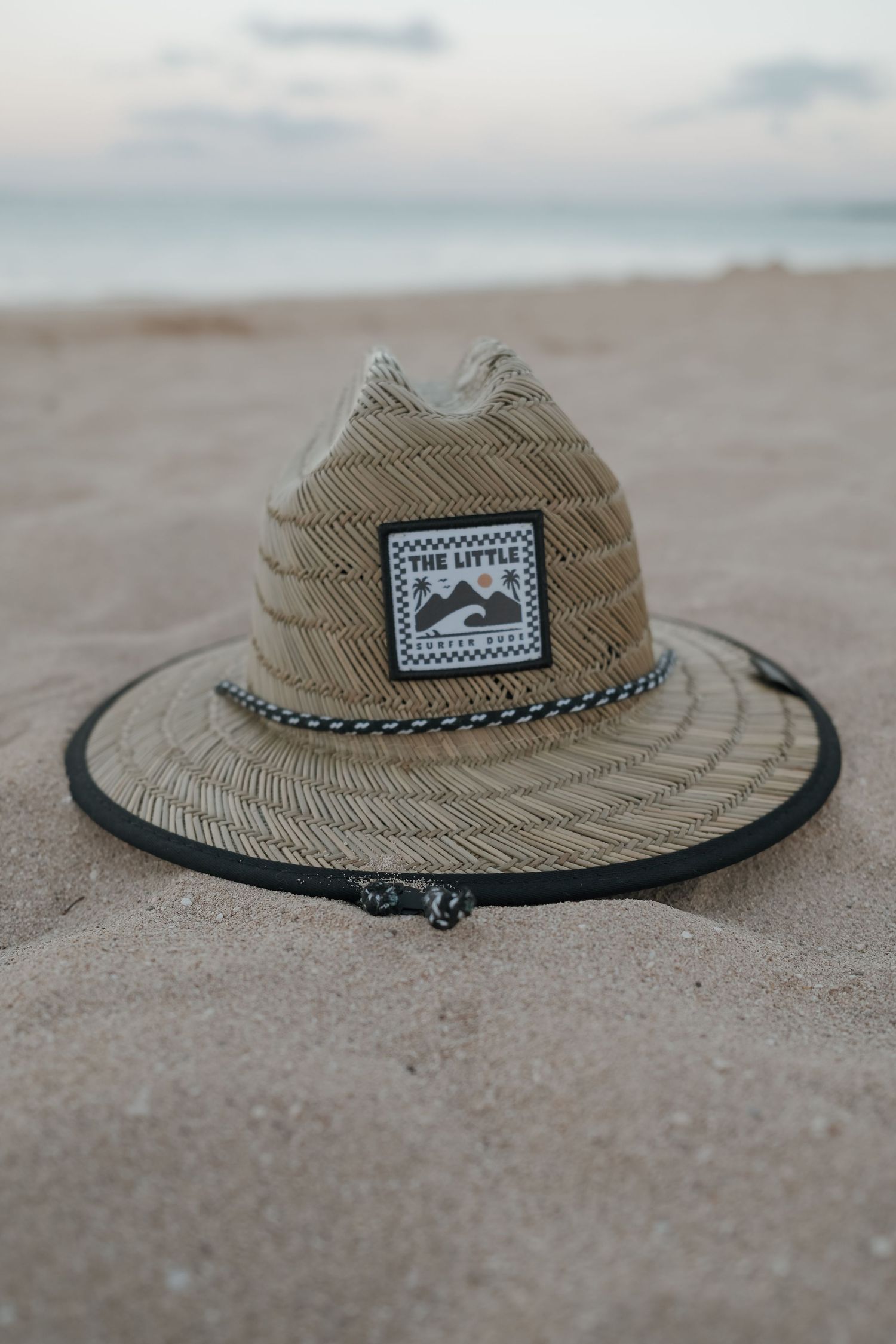 A straw lifeguard hat with a mountain patch and black rope trim resting on sandy beach at dusk.