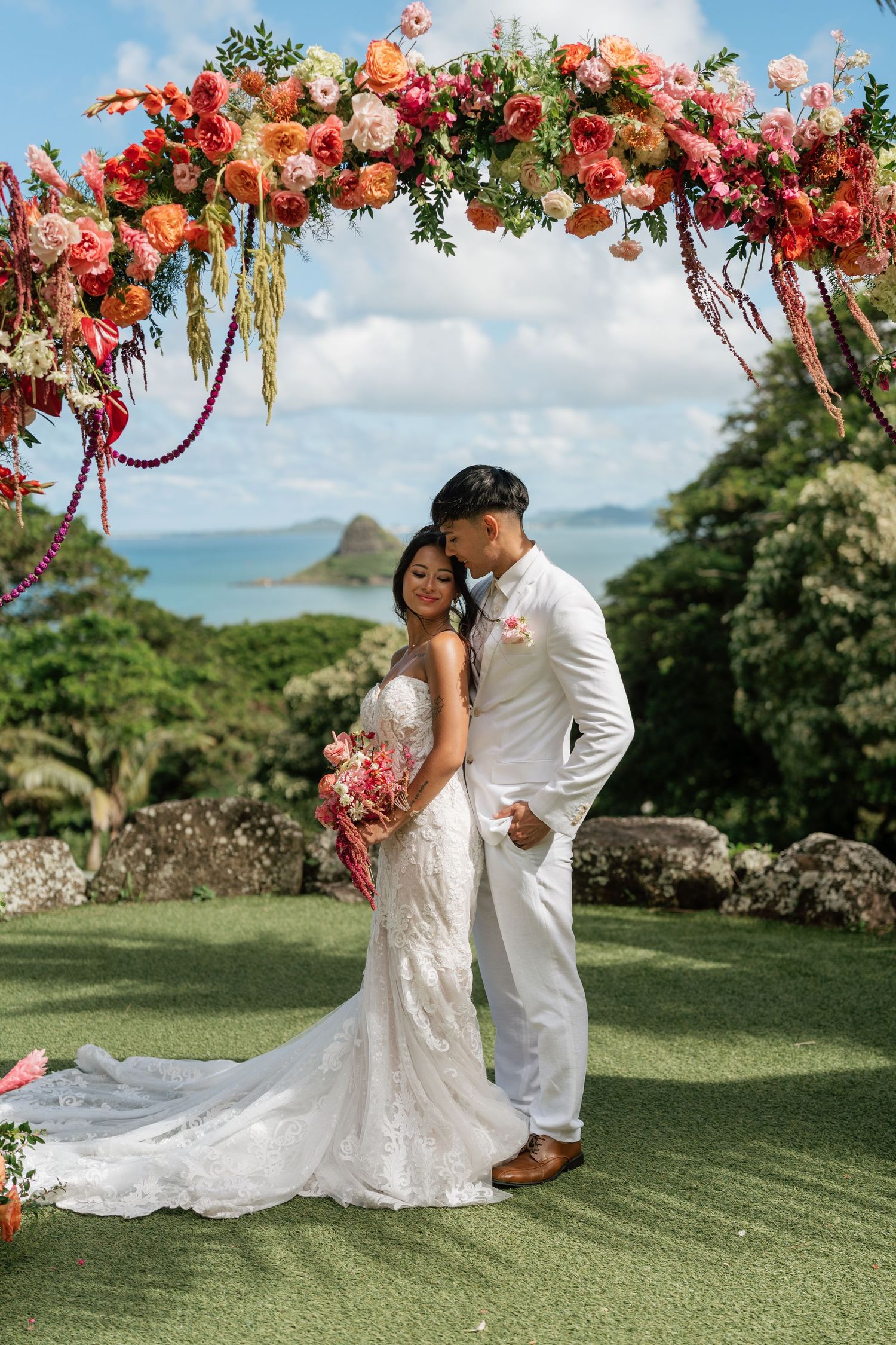 Couple poses under vibrant floral arch with ocean views at Hawaiian outdoor wedding ceremony.