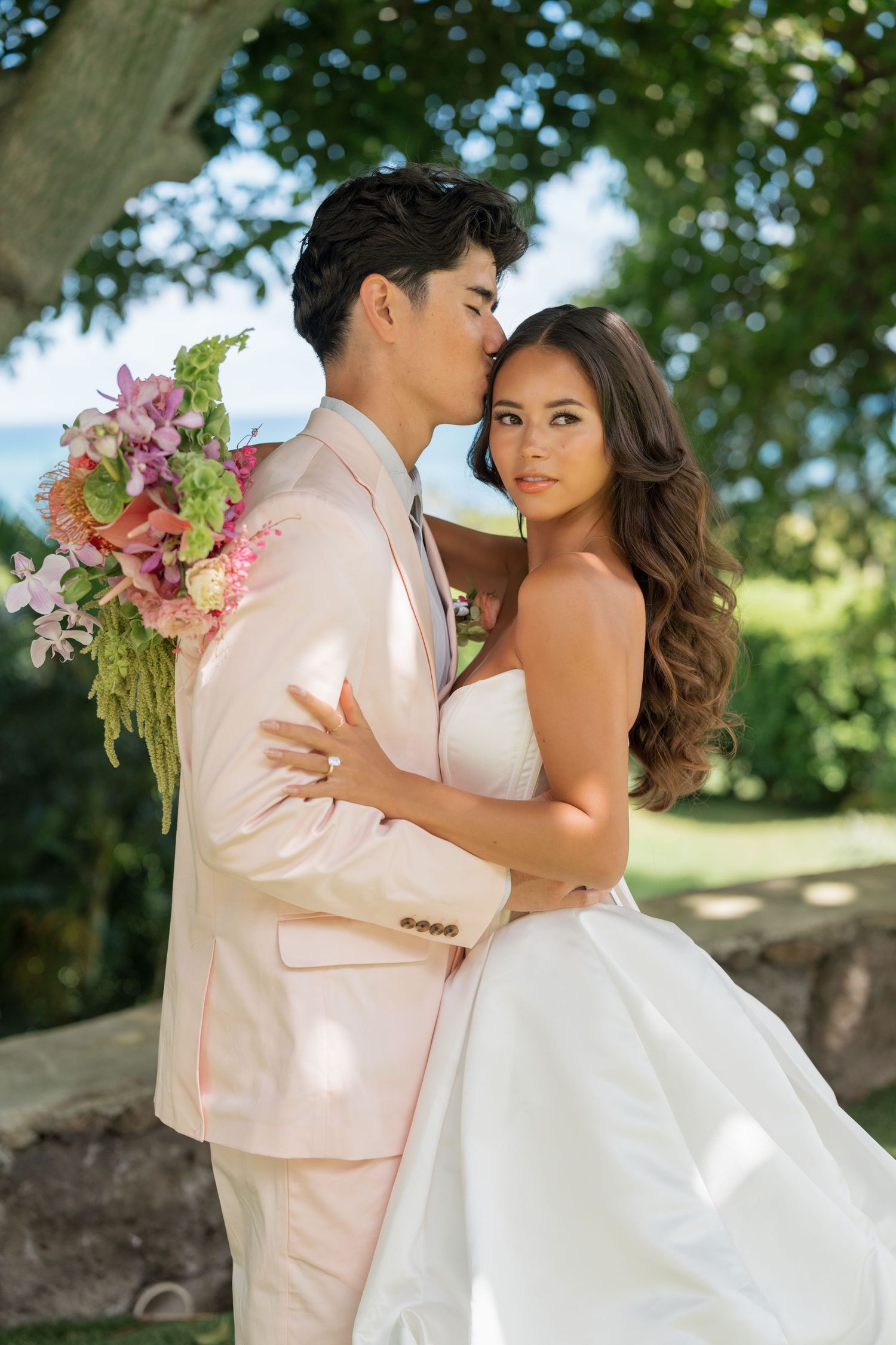 Bride in white strapless gown embraces groom in blush pink suit holding pink floral bouquet outdoors.