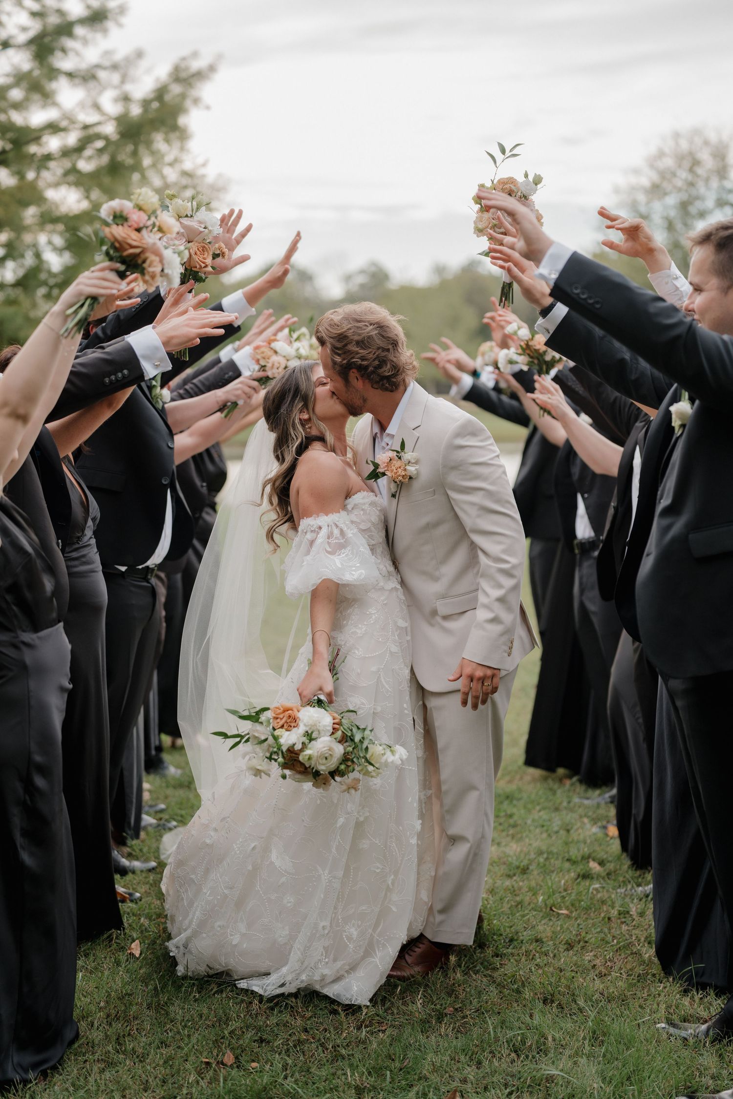 A romantic wedding moment as a couple shares a kiss while guests in dark attire raise their hands in celebration.