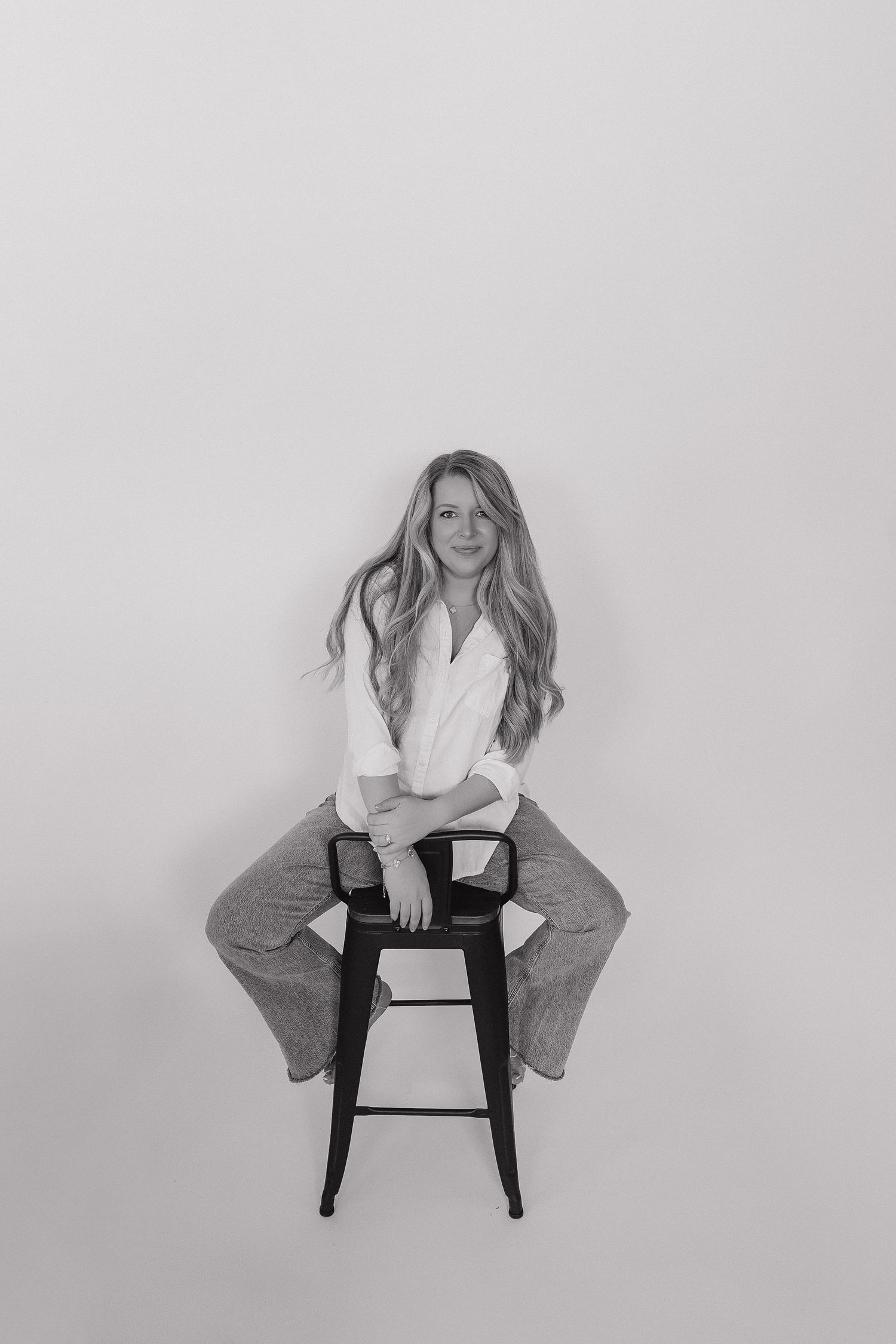 Black and white portrait of a young woman with long hair sitting on a stool, wearing a white shirt and jeans.