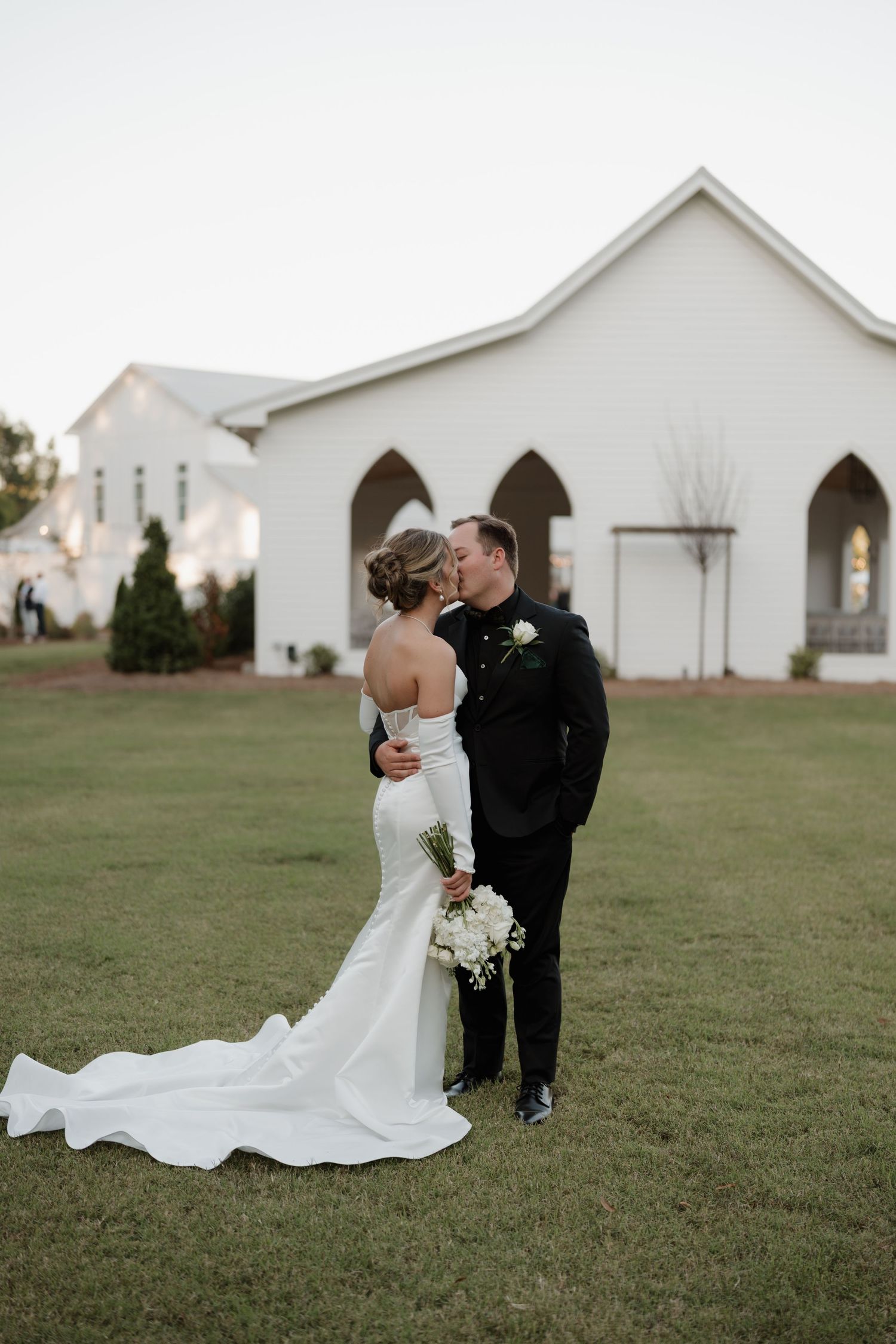 A couple shares a romantic moment on a grassy lawn with white church buildings in the background.