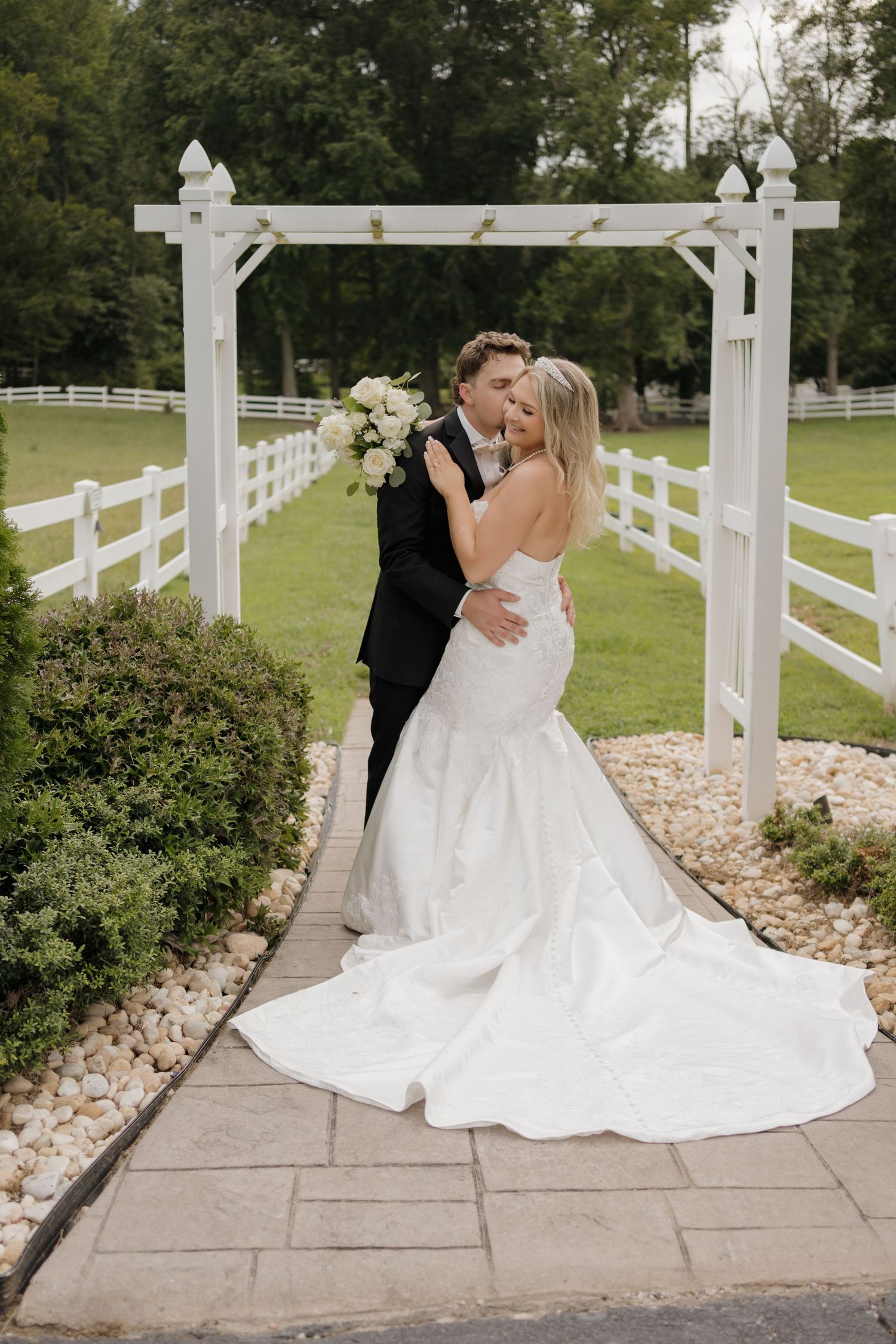 A romantic wedding couple embraces near a white fence pergola in an outdoor garden setting with lush greenery.