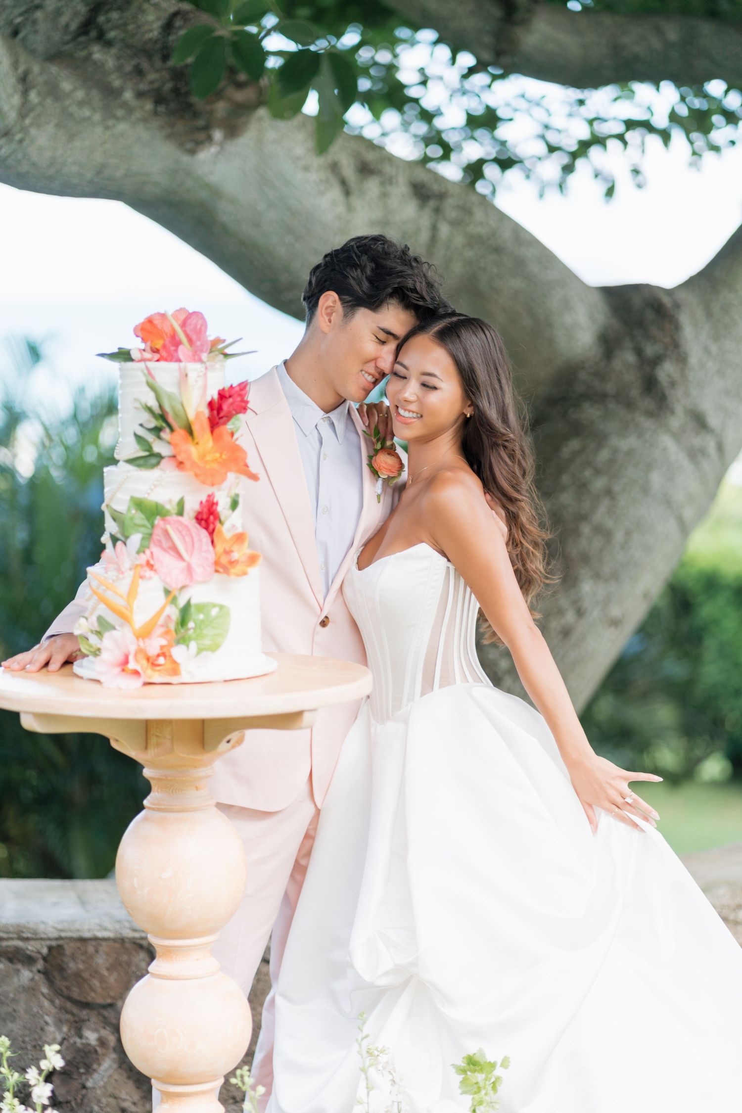 Bride and groom pose beside a floral wedding cake under a tree at an outdoor tropical wedding ceremony.