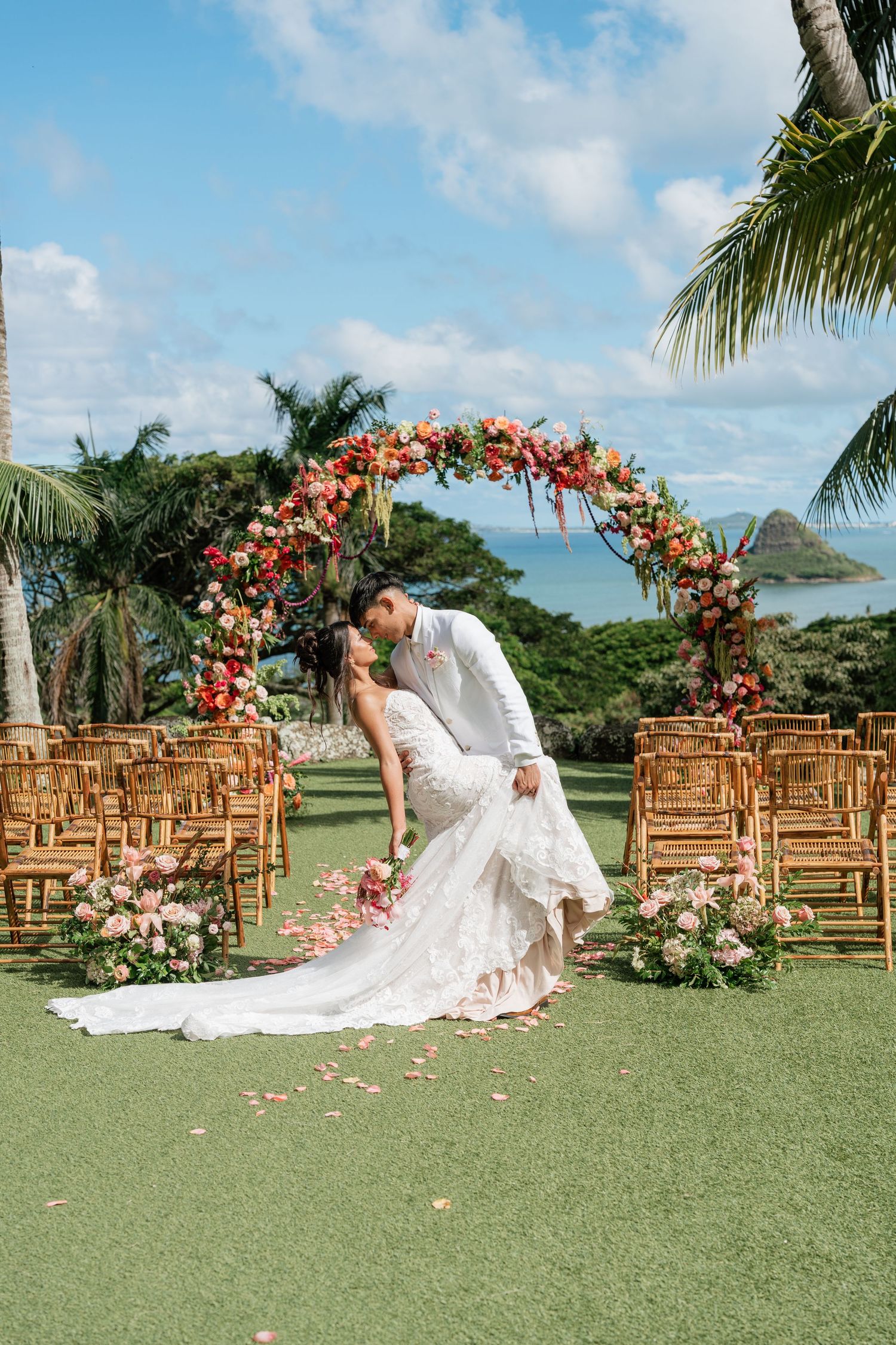Bride and groom share a romantic dip kiss at an outdoor Hawaii wedding ceremony with a floral arch and ocean views.