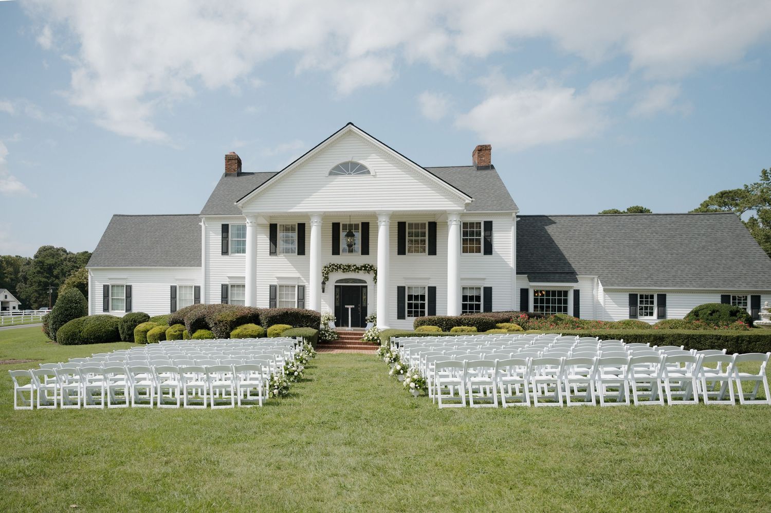White wedding chairs arranged in neat rows on a green lawn facing an elegant Southern colonial style house.
