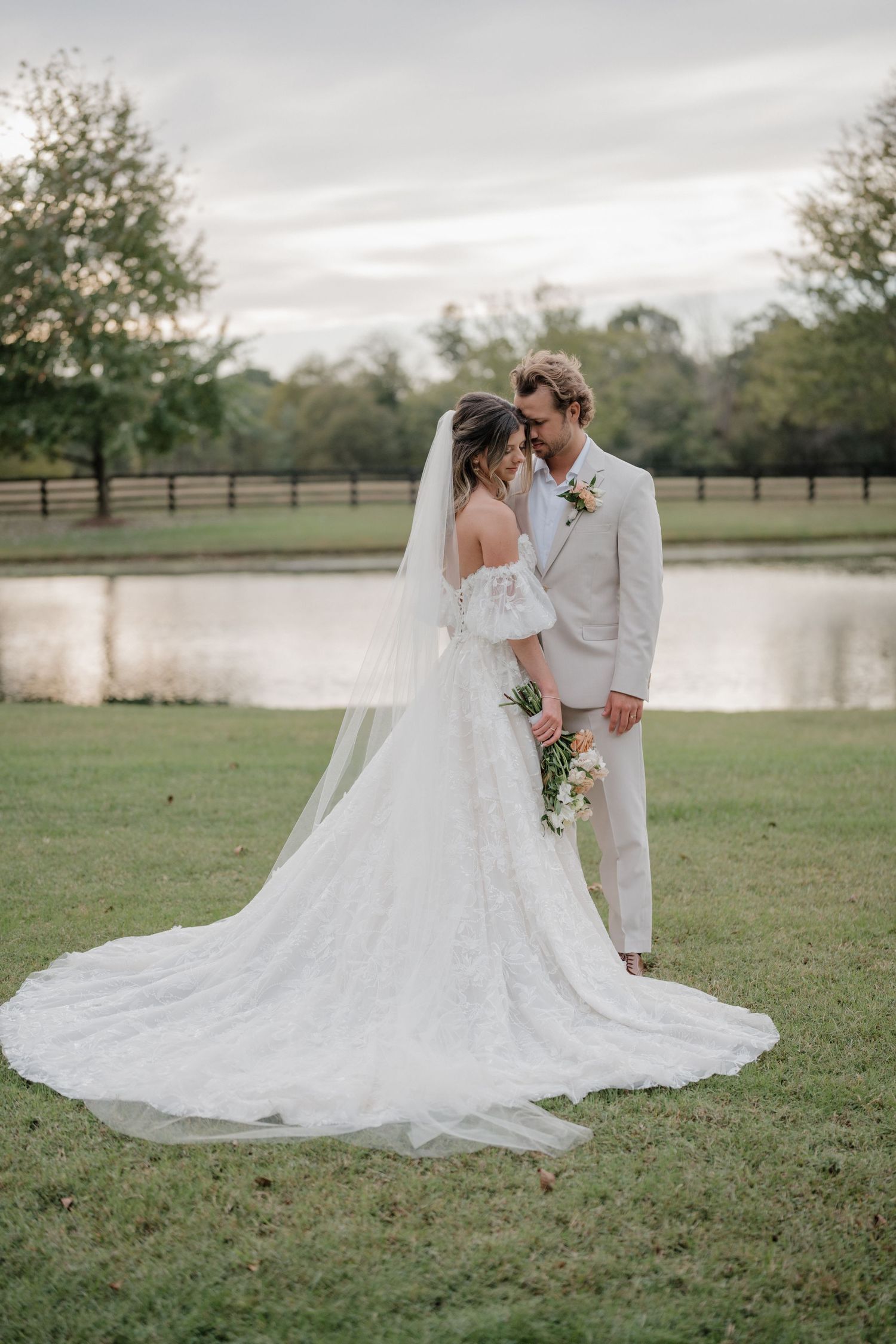 A romantic wedding couple embraces on a grassy field near a serene lake during their outdoor countryside celebration.