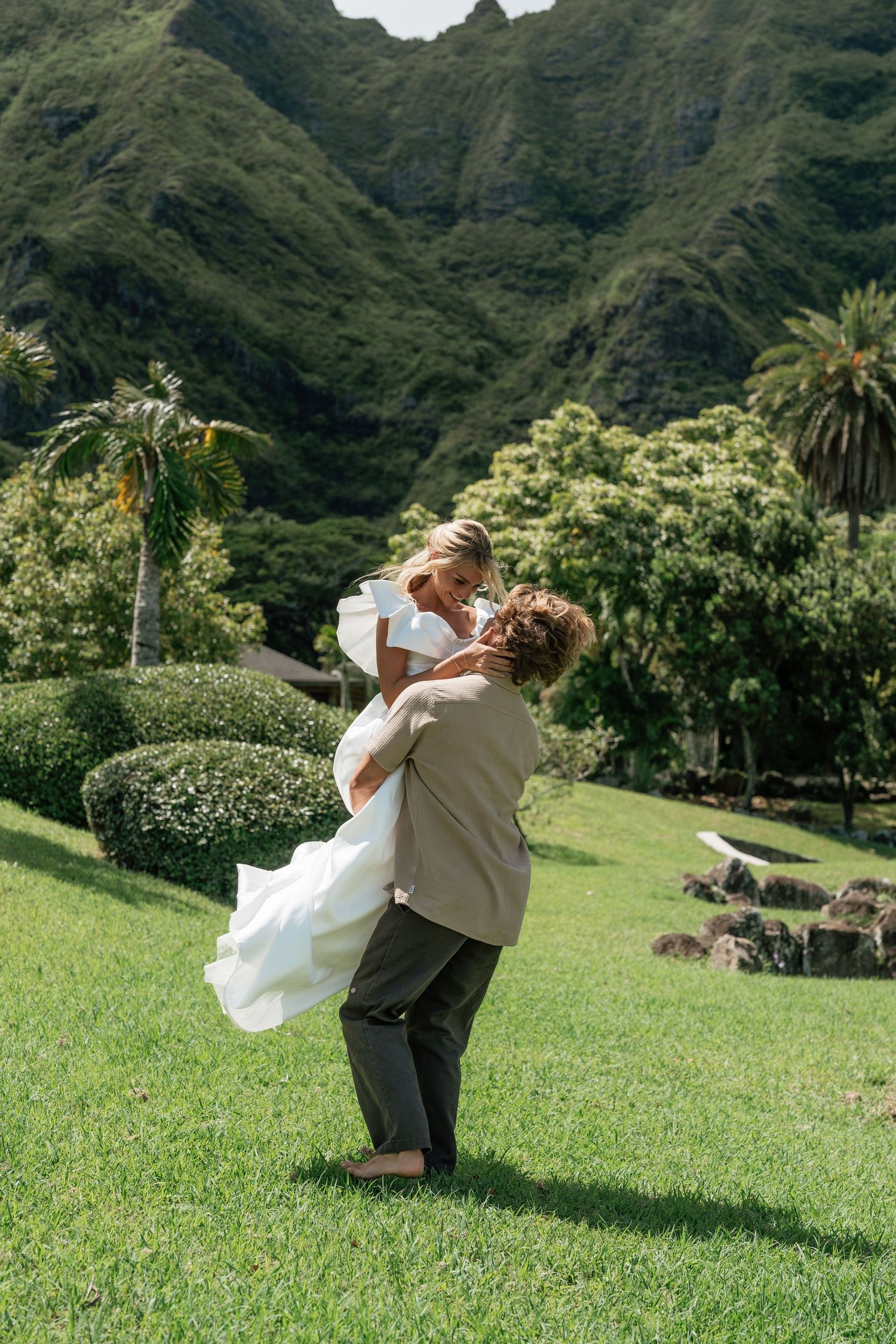 Groom lifting bride in white dress on lush green lawn with tropical mountain backdrop at Hawaii wedding.