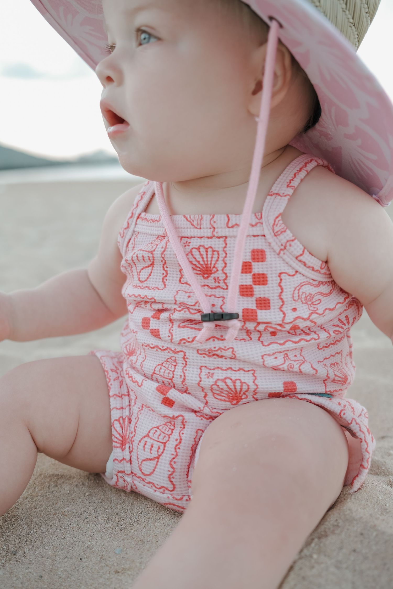 Baby girl in pink seashell print romper and sun hat sitting on sandy beach.