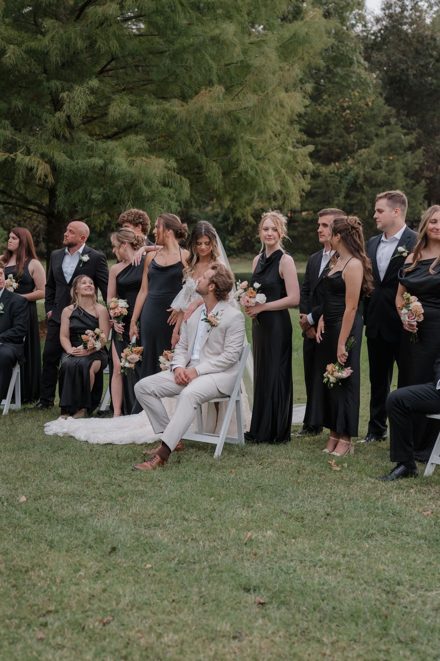 Wedding party in black dresses and grey suits gathered outdoors for a group photo on the lawn.