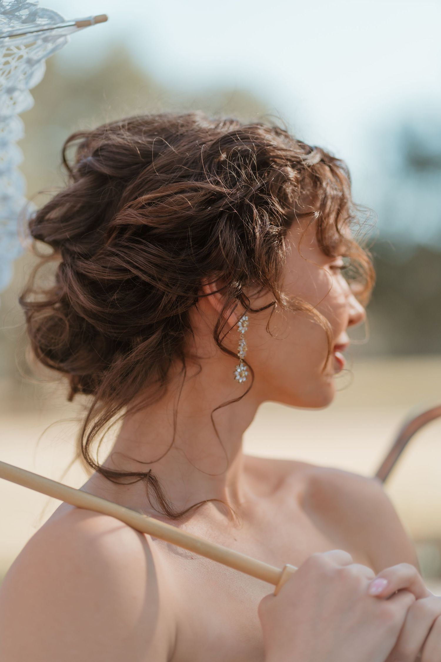 A bride with an elegant updo hairstyle holds a lace parasol outdoors, wearing delicate drop earrings.