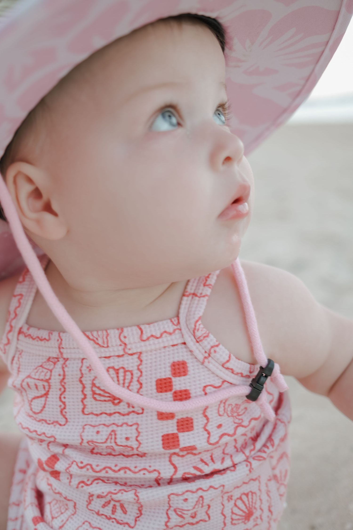 Adorable baby girl in pink sun hat and floral swimsuit looking up curiously at the beach.