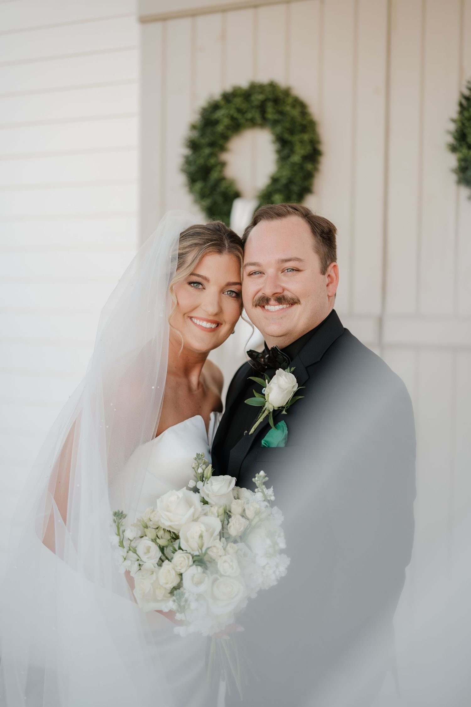 A couple poses on their wedding day in front of a white wall with an evergreen wreath, holding a white bouquet.