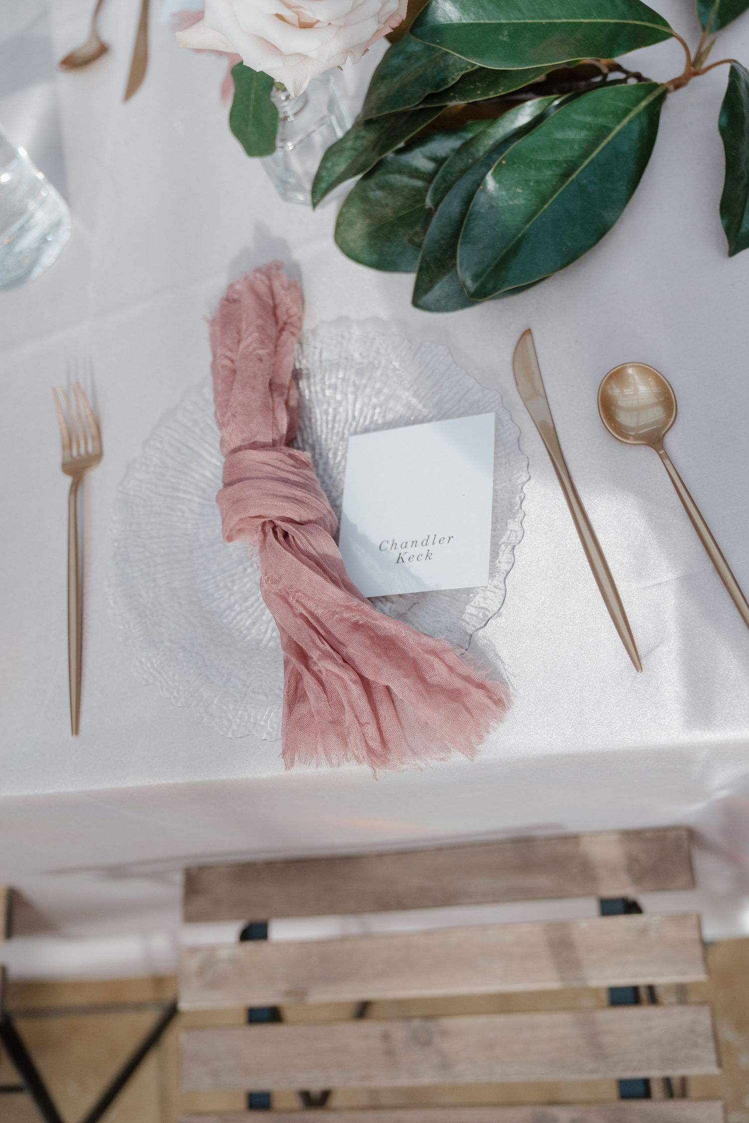Wedding table setting with gold utensils, magnolia leaves, and blush pink accents on white linens.