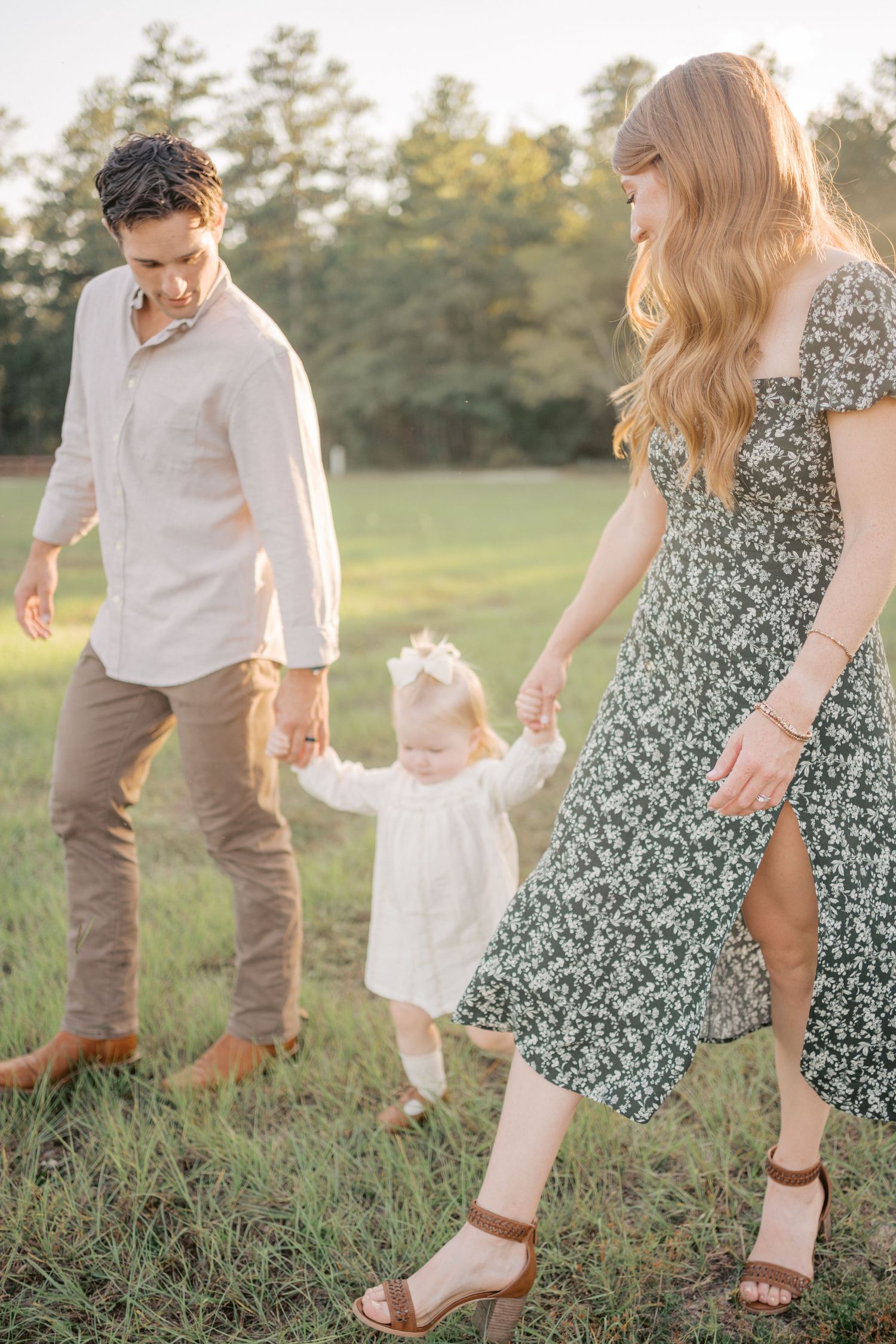 Family in casual attire walking together through a grassy field at sunset, helping small child learn to walk.