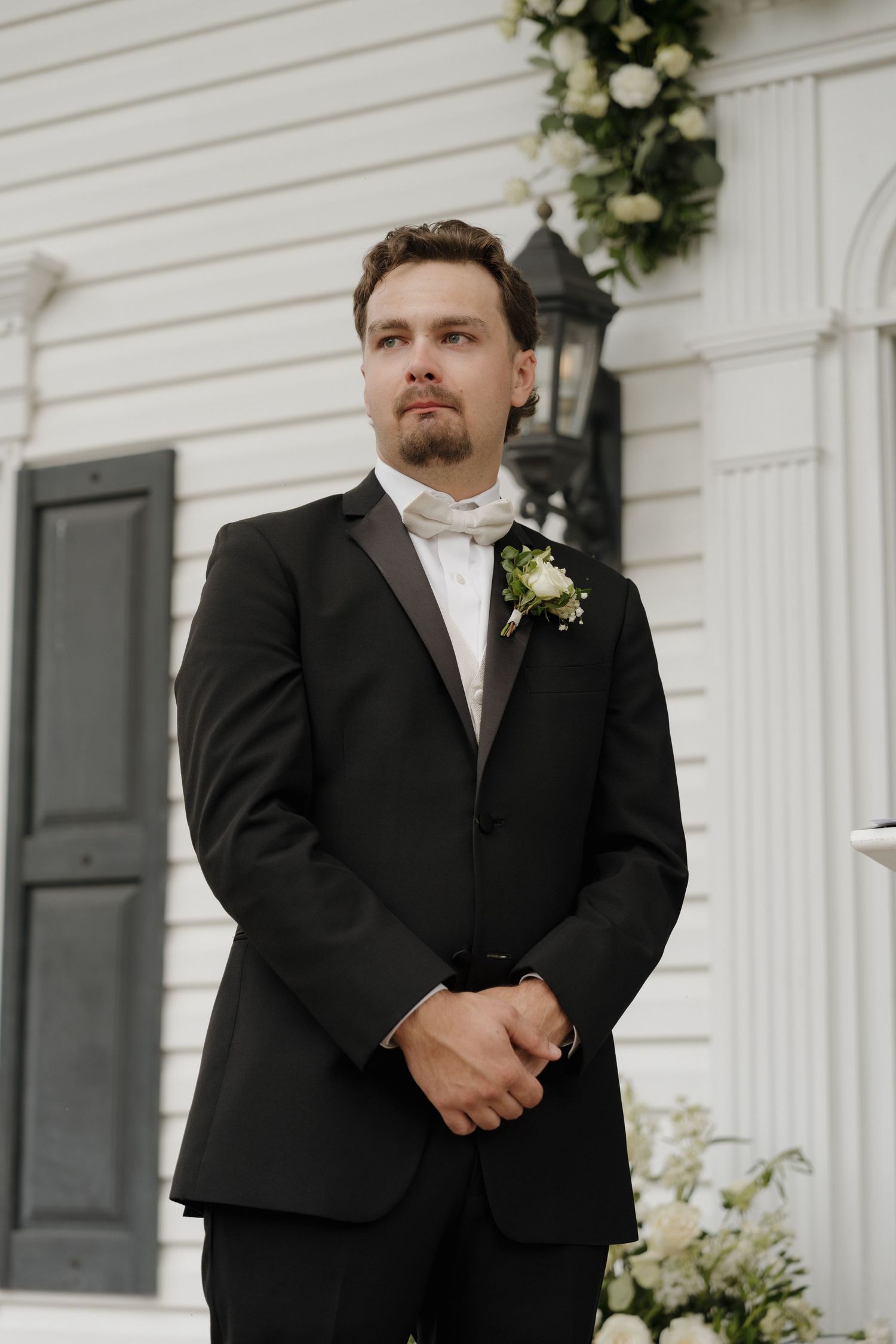 A groom in a black tuxedo and white bow tie stands outside against a white wall with floral decorations.