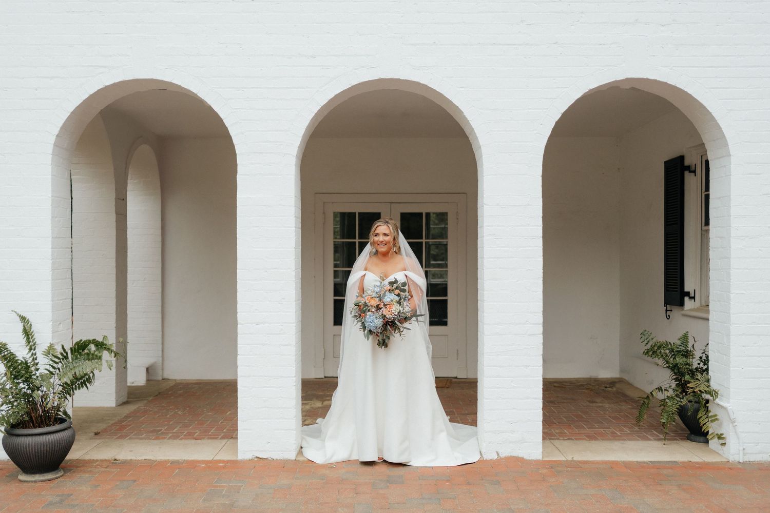 A beautiful white wedding dress is framed by elegant arched columns against a colonial-style white building exterior.