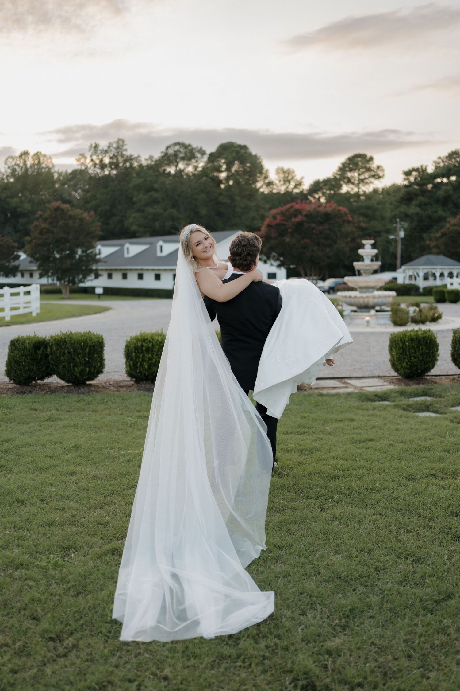 A romantic wedding couple embraces on a manicured lawn with a fountain and trees in the background at dusk.