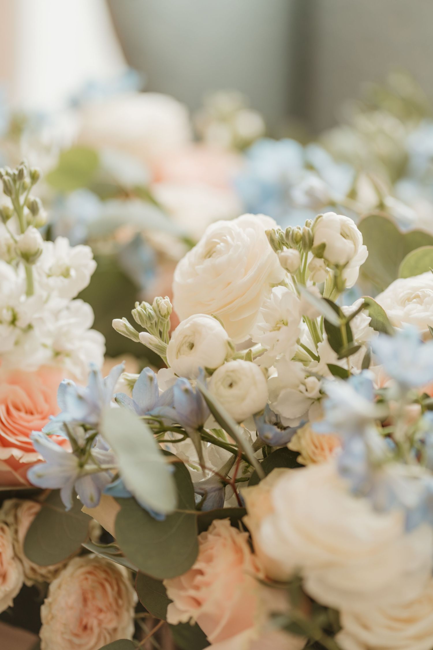 Elegant wedding bouquet with white ranunculus, peach roses, and blue hydrangeas arranged in a soft, romantic floral display.