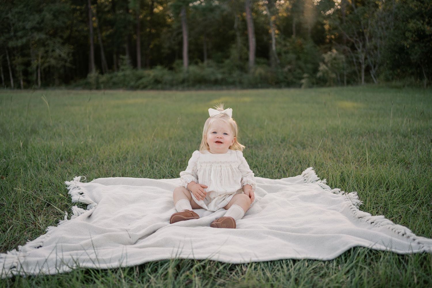 Small child wearing a cream colored dress sits on white blanket in grassy field at sunset.