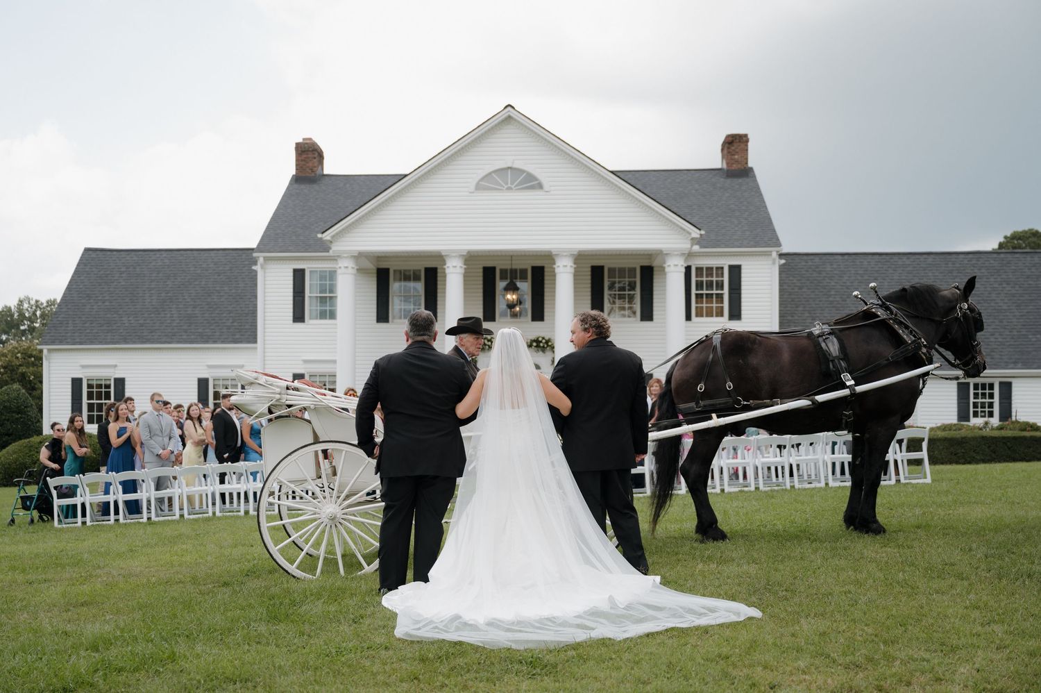 Wedding ceremony at a white farmhouse with guests seated outdoors on white chairs facing the colonial-style venue.