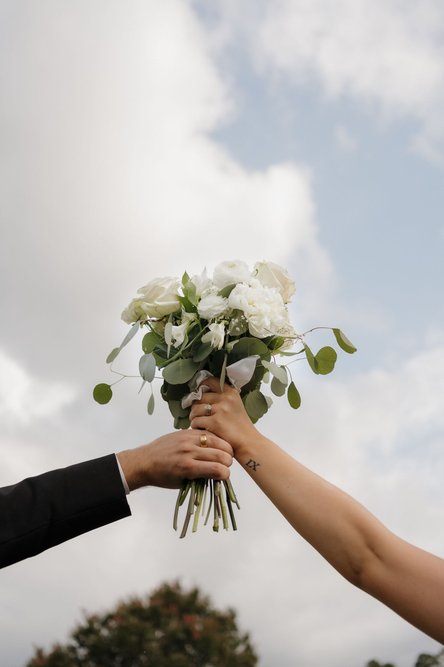 Two hands reaching toward each other with a white rose bouquet against a cloudy sky backdrop.