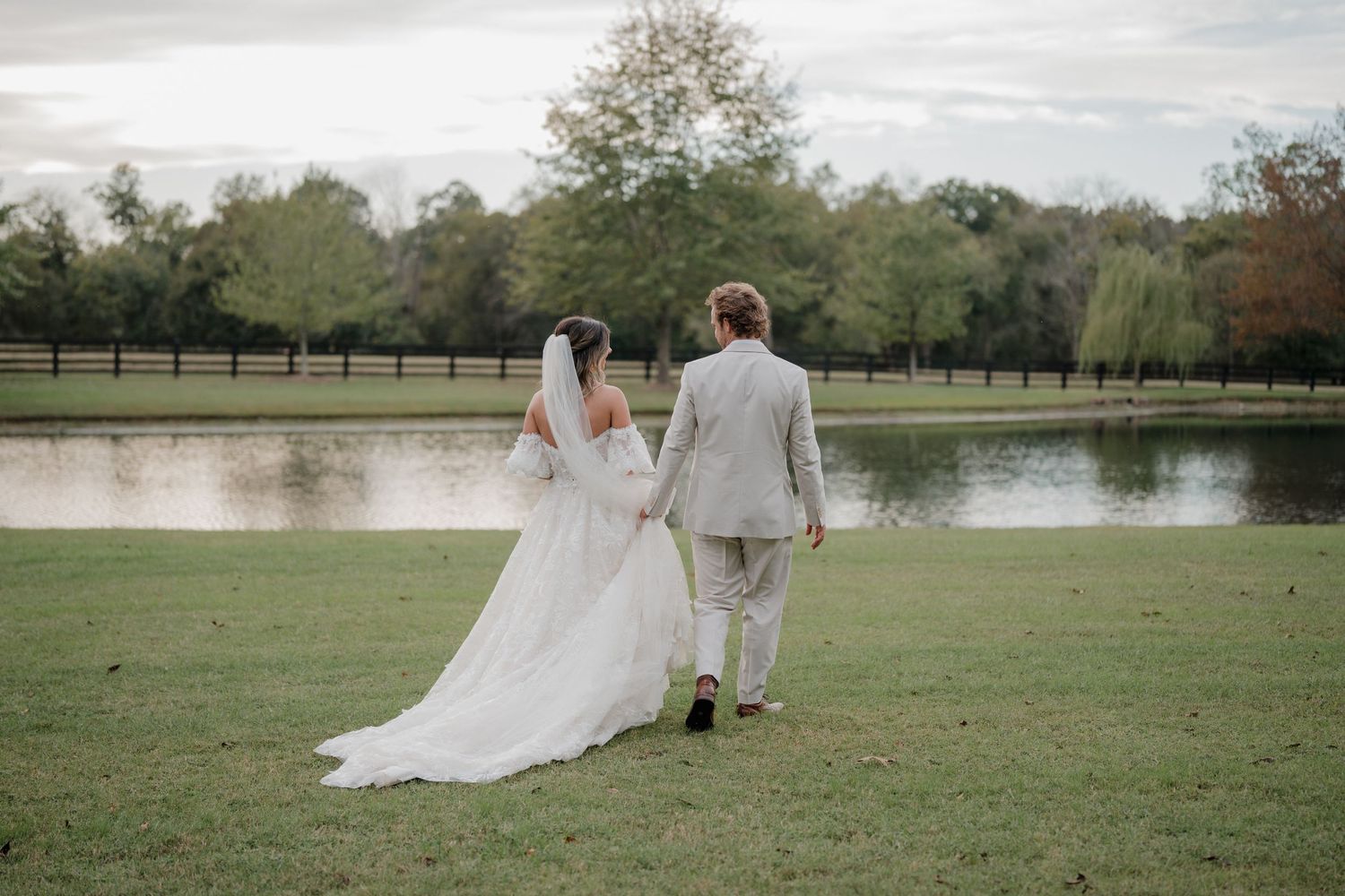 Newlywed couple walks together through a scenic park setting with a lake and trees in the background.