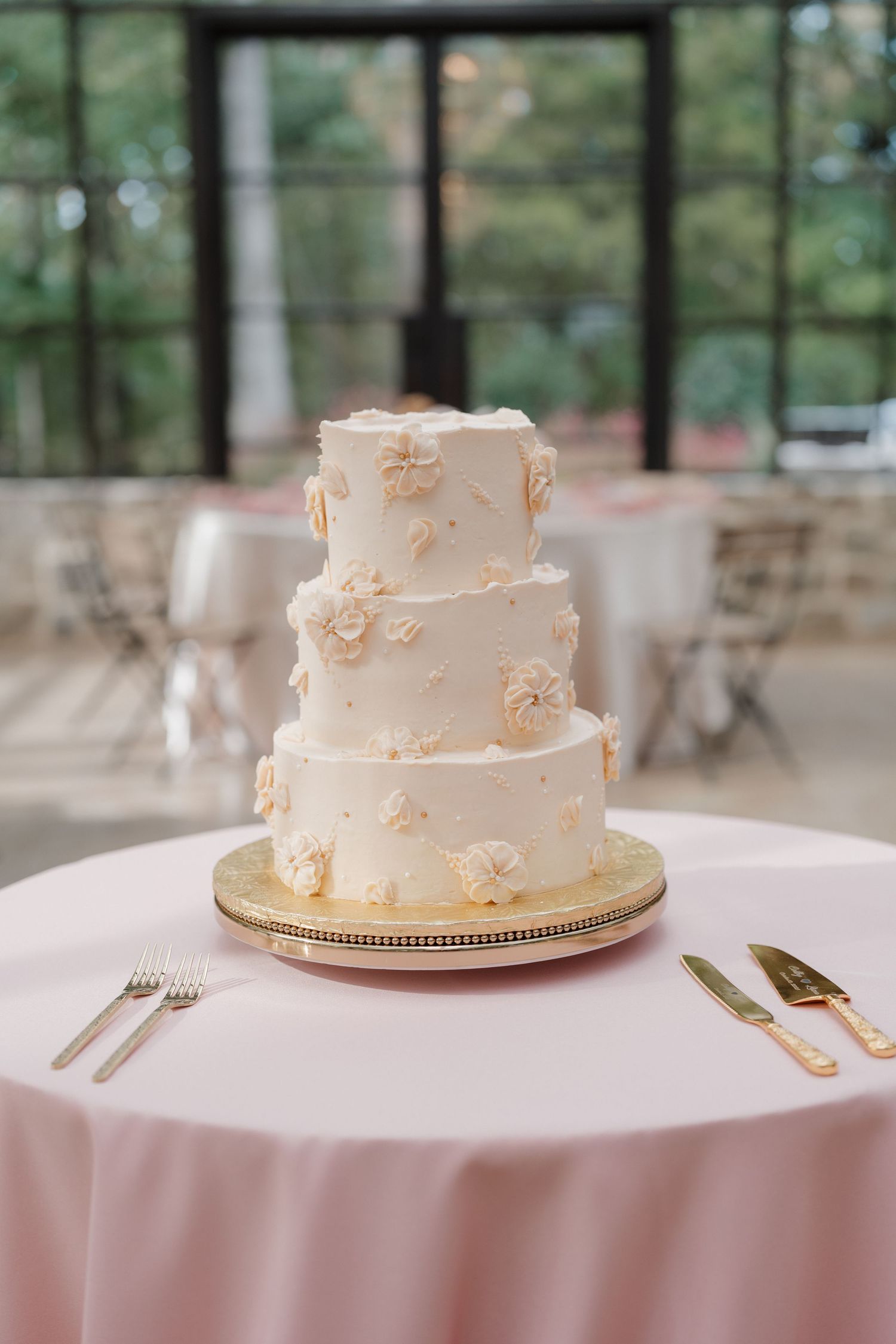Elegant three-tier white wedding cake with delicate floral details displayed on a pink tablecloth with gold cake stand.