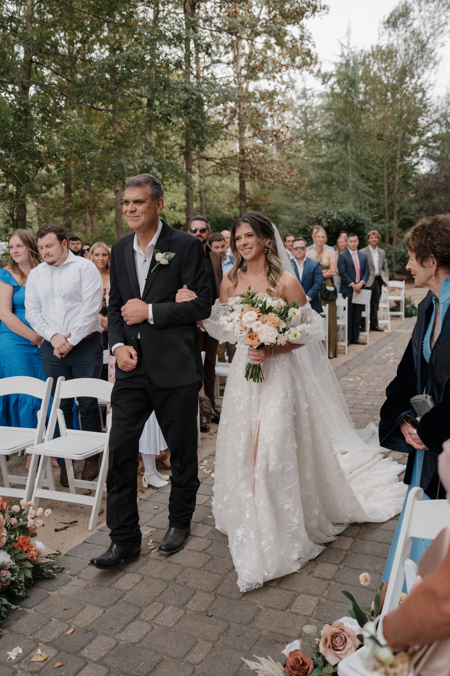Wedding ceremony processional on stone path with guests seated on white chairs outdoors.