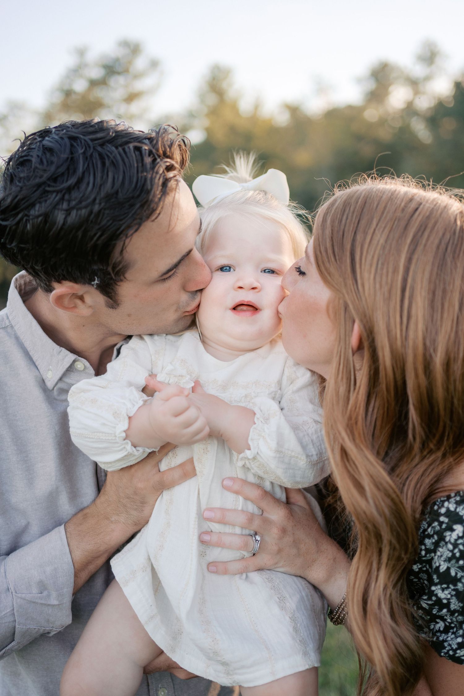 A tender family moment captures affectionate parents kissing their baby in a white dress during a sunny outdoor photoshoot.