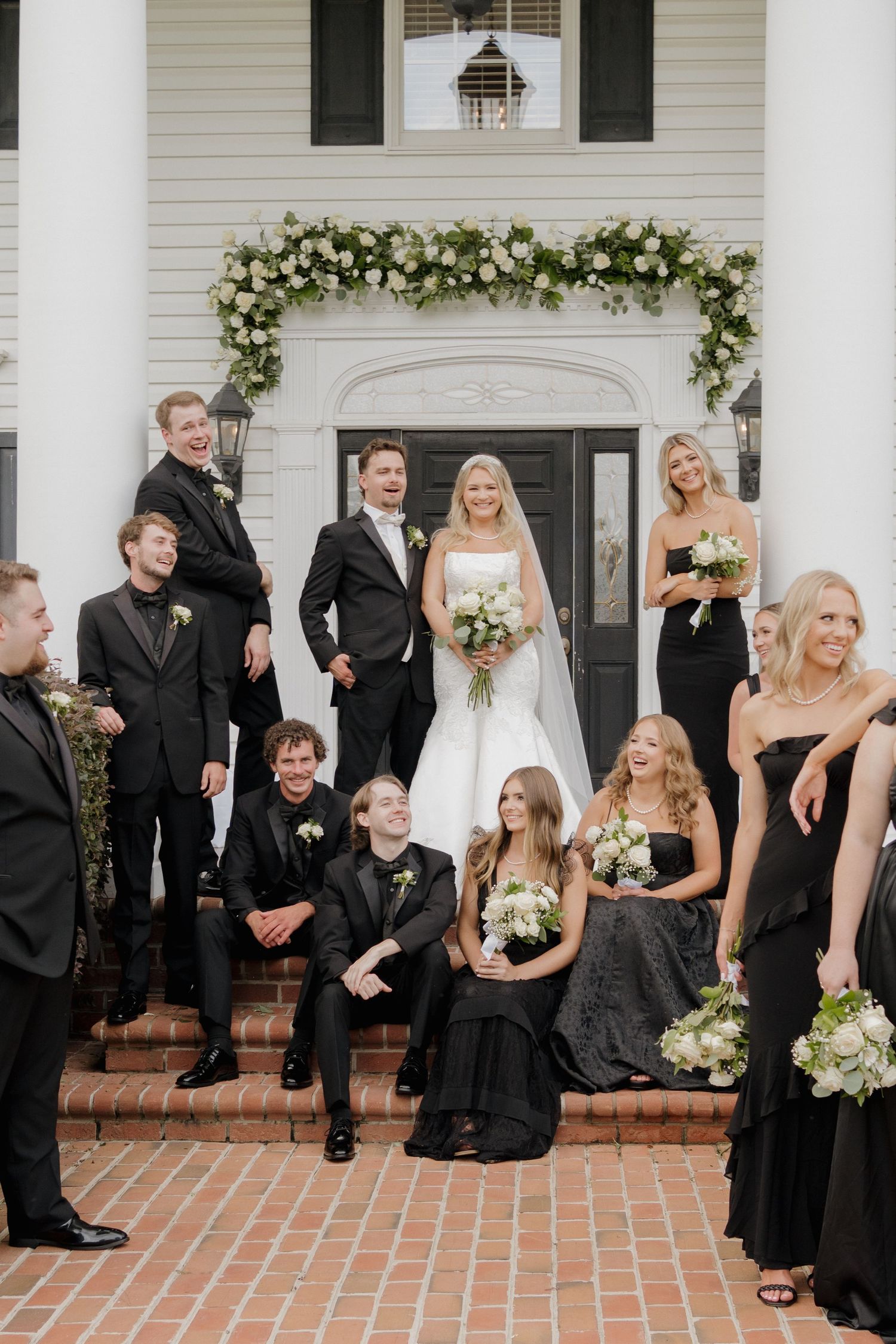 Wedding party in black attire laughs together during formal photos on brick steps of white house with floral arch.