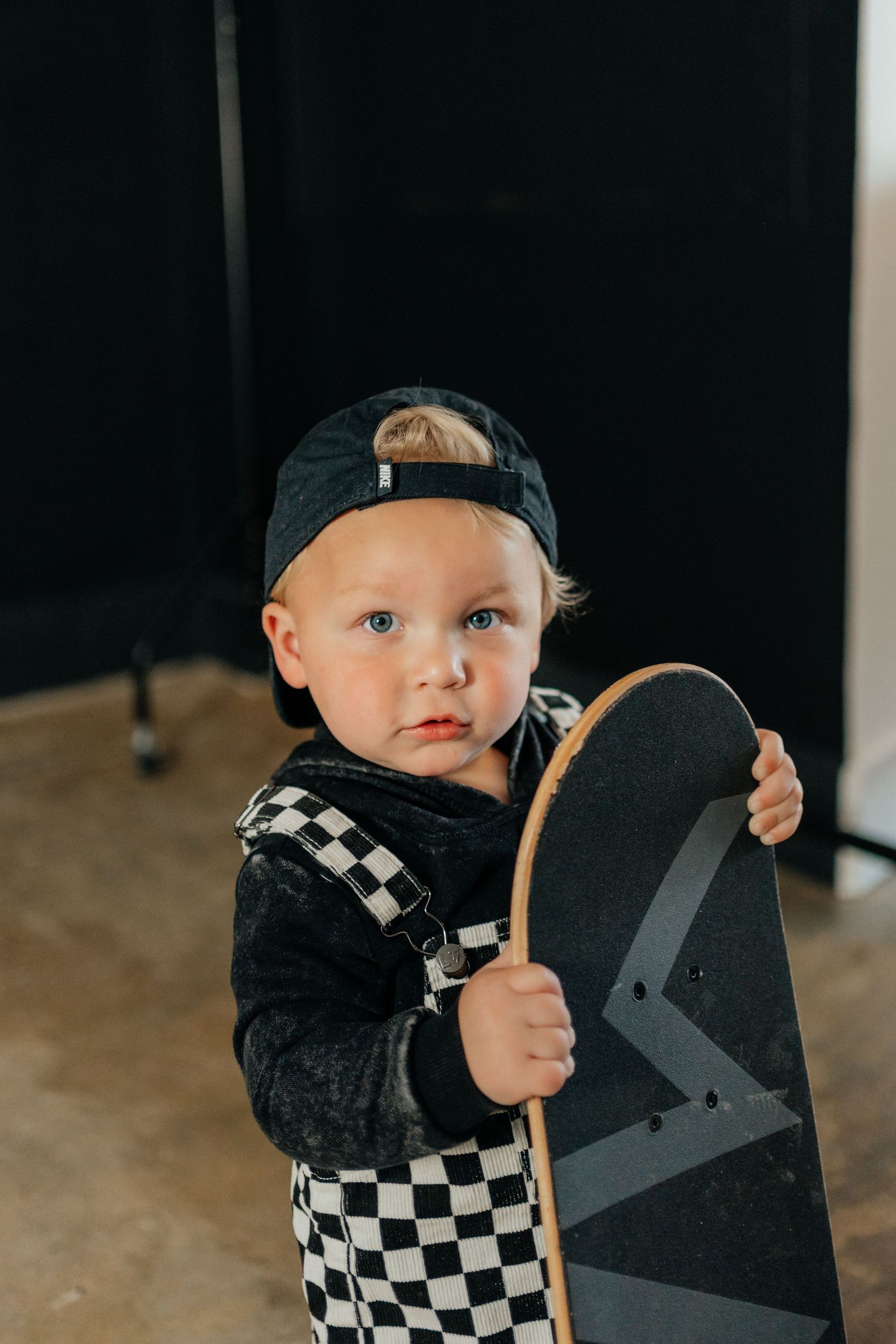 Cute toddler boy in checkered pants and snapback cap holding a skateboard indoors.