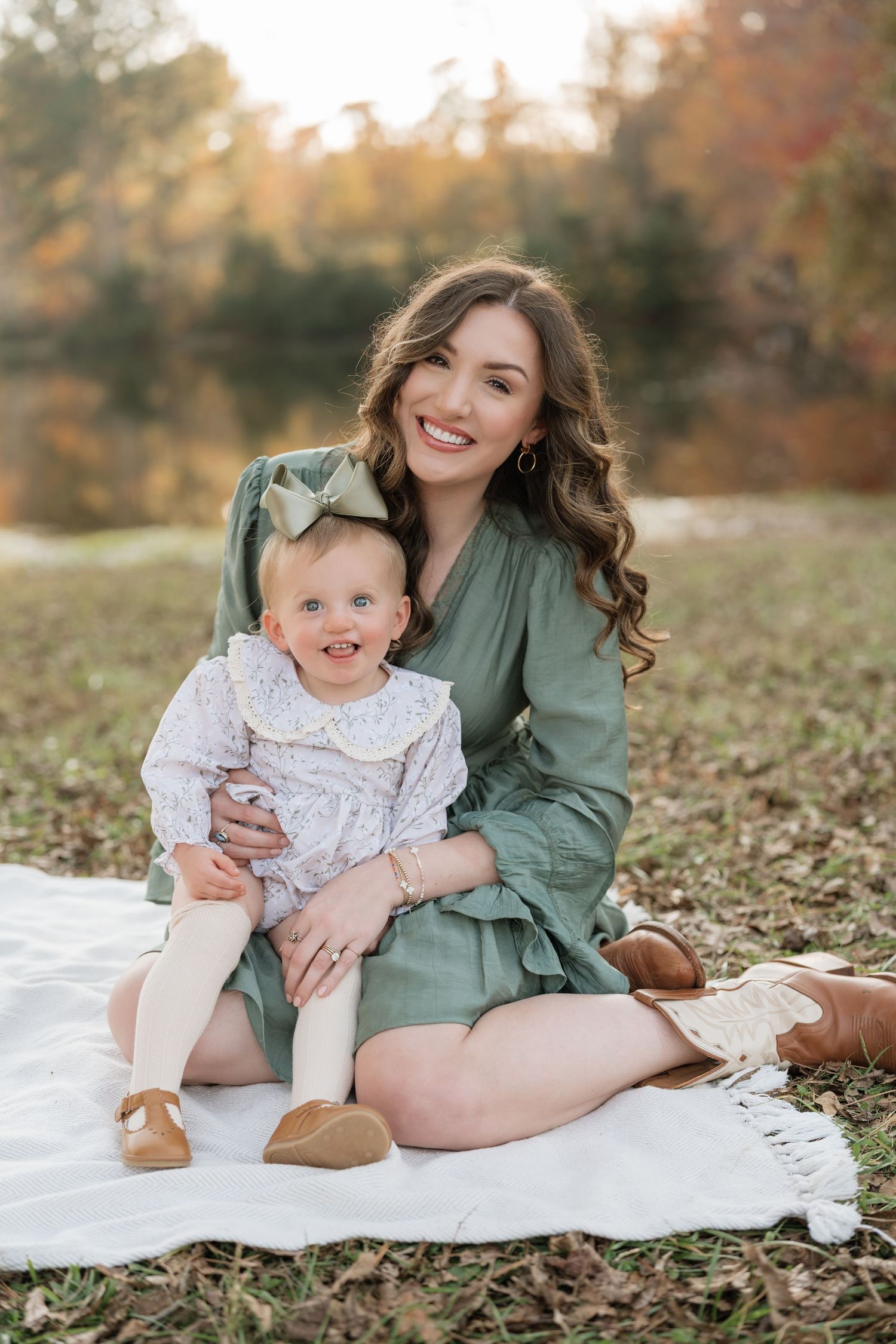 Mother and child sit together on white blanket during autumn photoshoot outdoors among fallen leaves.