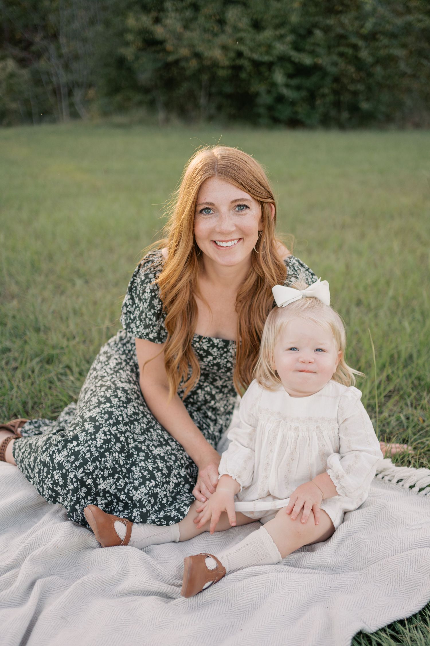 A parent and child sit together on a blanket in a grassy field during a golden hour photoshoot.