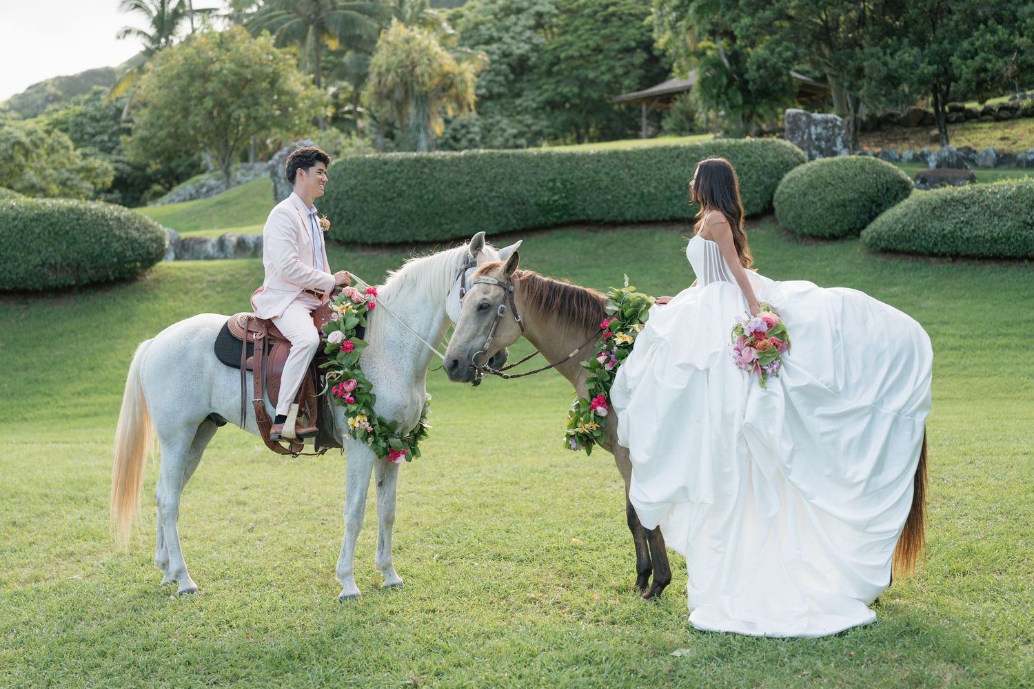 Bride and groom pose with flower-adorned horses on lush green lawn in elegant outdoor wedding photo.